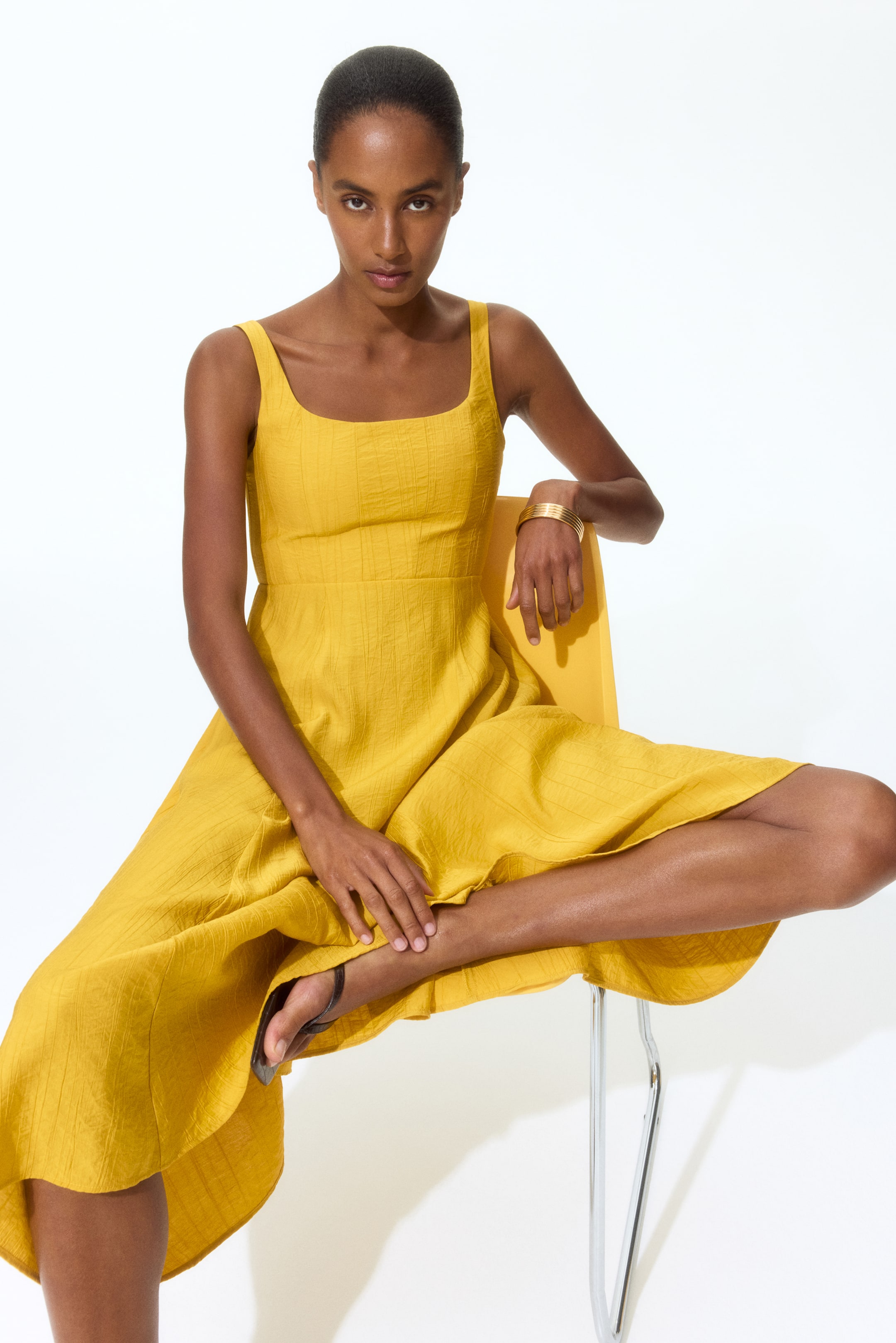 View larger image: The yellow dress showcases a square neckline paired with broad shoulder straps. The fitted bodice gracefully transitions into a voluminous skirt crafted from crinkled fabric, adorned with a subtle grid pattern.