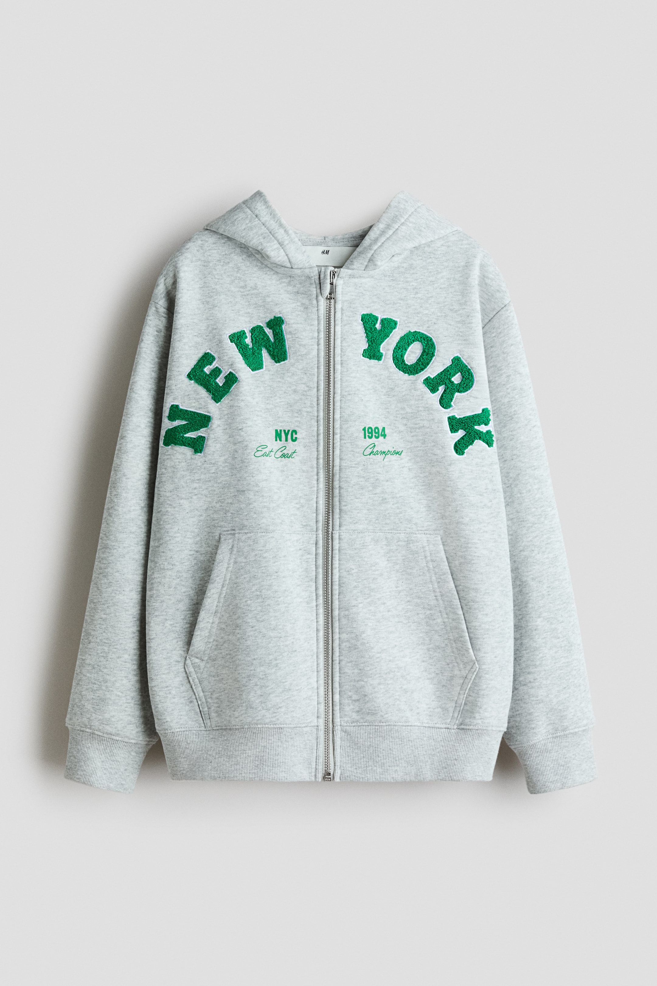 View larger image: A grey full-zip hoodie is presenting FRONT, featuring an attached hood, ribbed cuffs and hem, and a kangaroo pocket. The chest is displaying arched, textured green lettering spelling "NEW YORK," with smaller embroidered text "NYC East Coast" and "1994 Champions" positioned below.