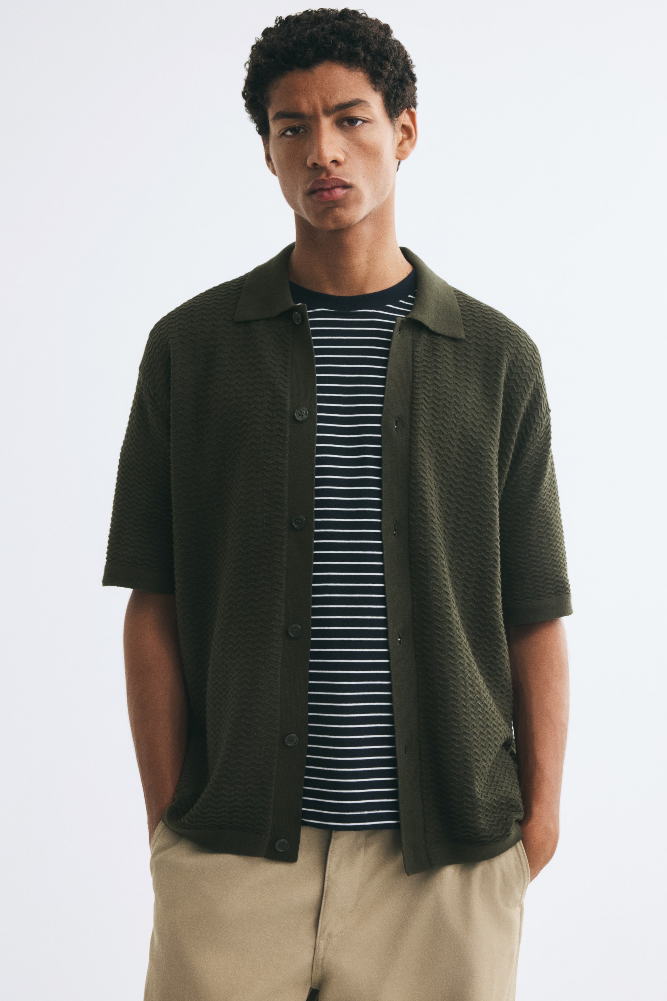 View larger image: A dark green, textured-knit short-sleeve shirt is worn unbuttoned by the model, featuring a flat collar, button-down front, and fine ribbing at the cuffs and hem. The shirt is layered over a dark blue and white horizontally striped t-shirt and paired with beige trousers.