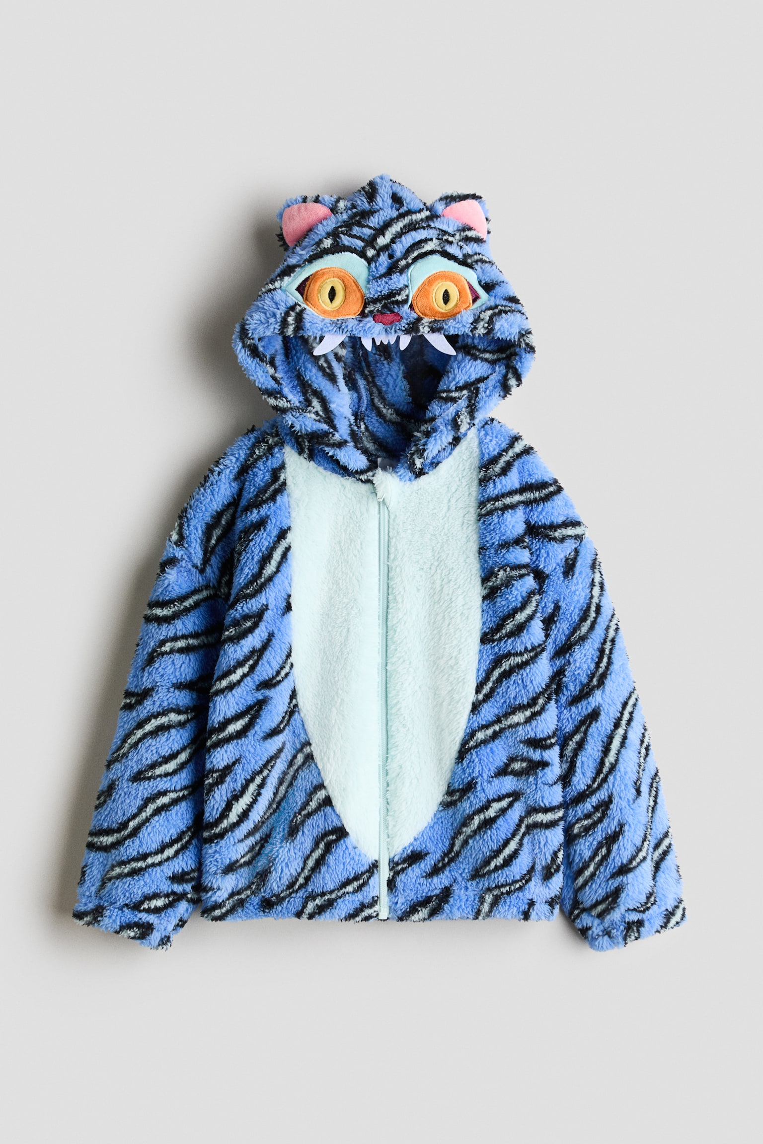 A blue and black tiger-striped fleece zip-up hoodie is shown from the front, featuring a light blue contrast panel on the chest. The hood displays an embroidered monster face with large orange eyes, small pink ears, and white felt fangs hanging along the edge.