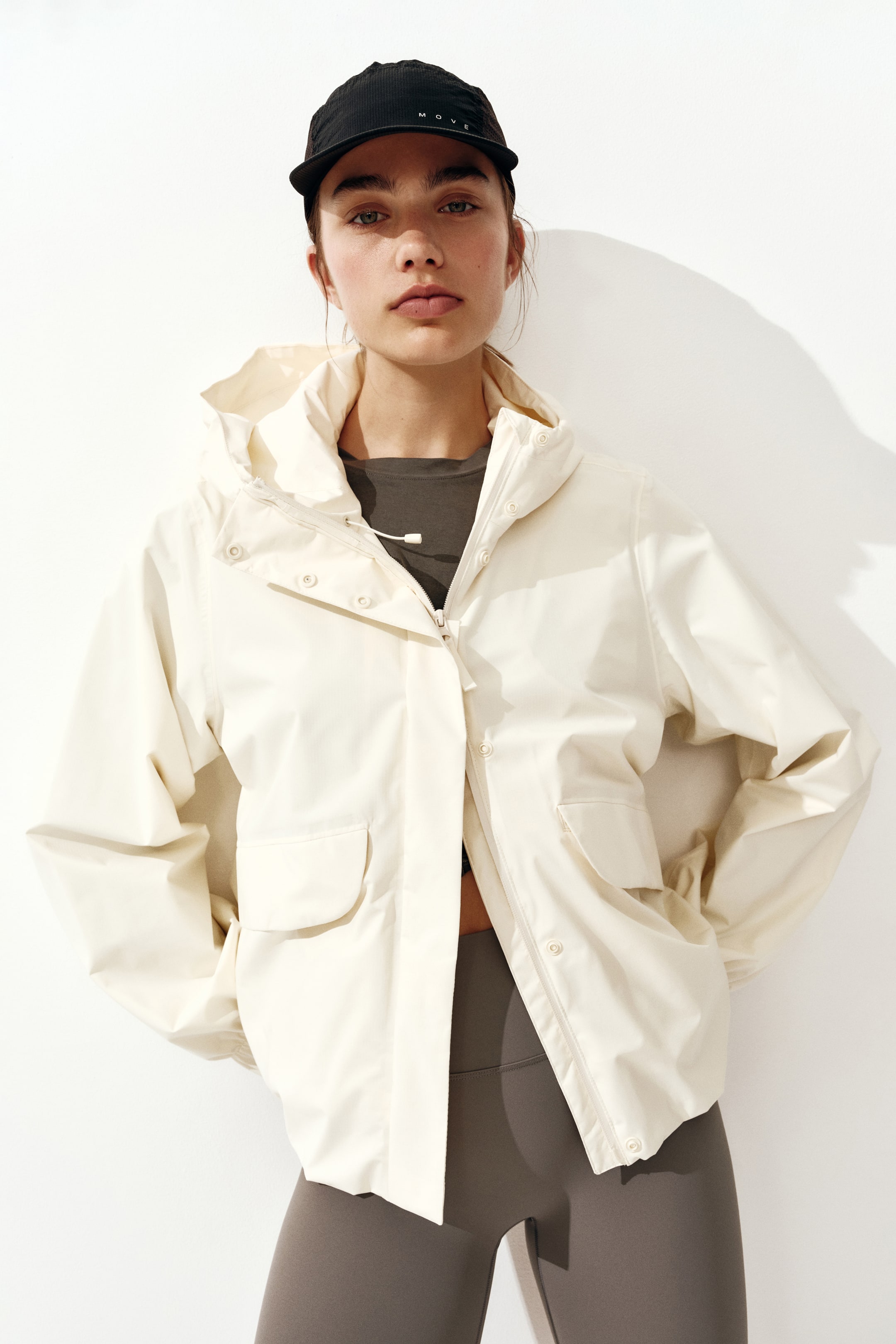 View larger image: An off-white hooded jacket, crafted from a subtly textured fabric, is presented slightly angled to the left. It features a partial zip and snap-button placket closure, a high collar with a drawstring, an attached hood, and two large front flap pockets. It is worn open over a dark top, with dark leggings visible below its cropped hem.