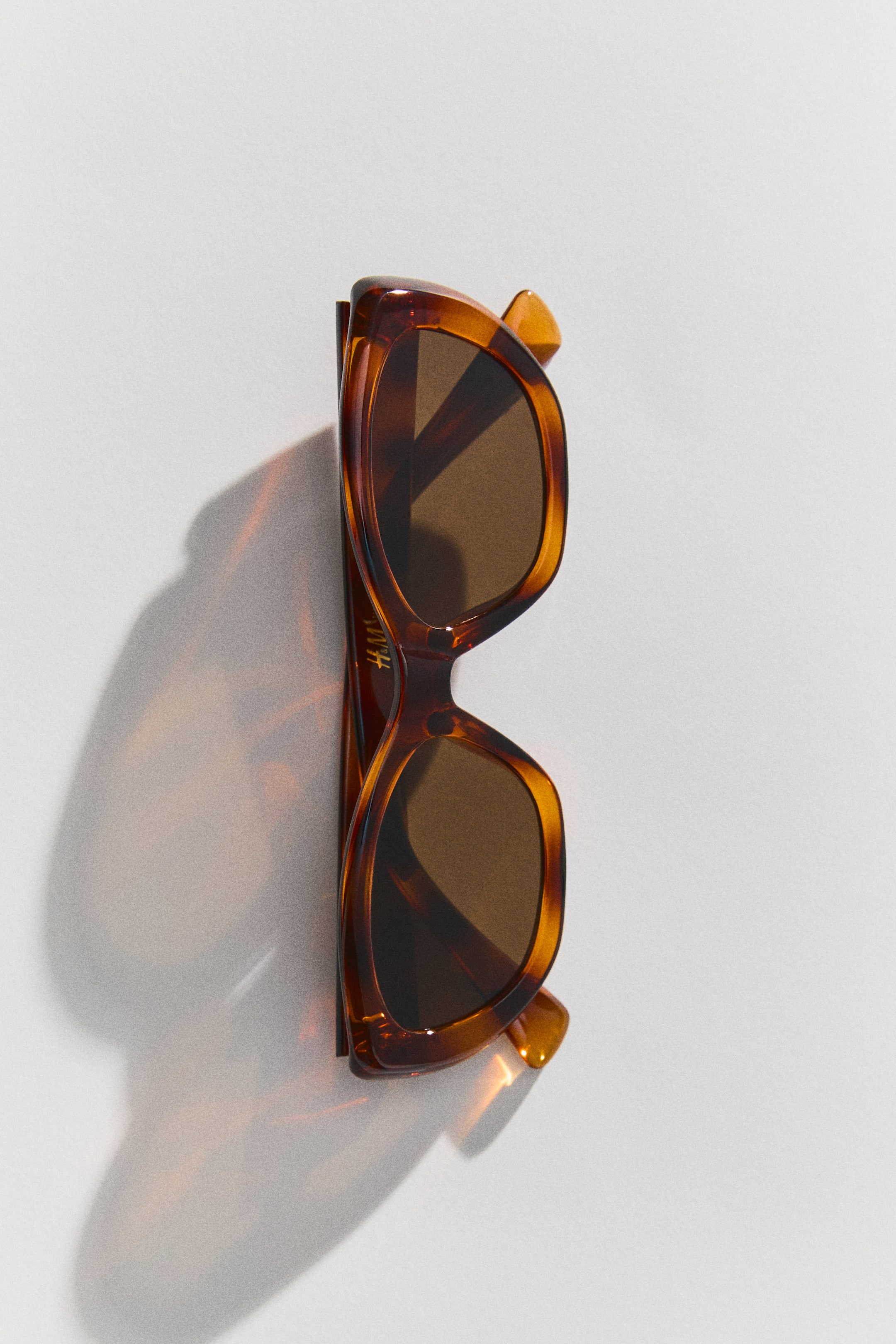View larger image: Cat-eye sunglasses with tortoiseshell-patterned plastic frames and dark brown tinted lenses are resting upright, shown from the front. The sunglasses cast a warm, translucent amber shadow to the side.