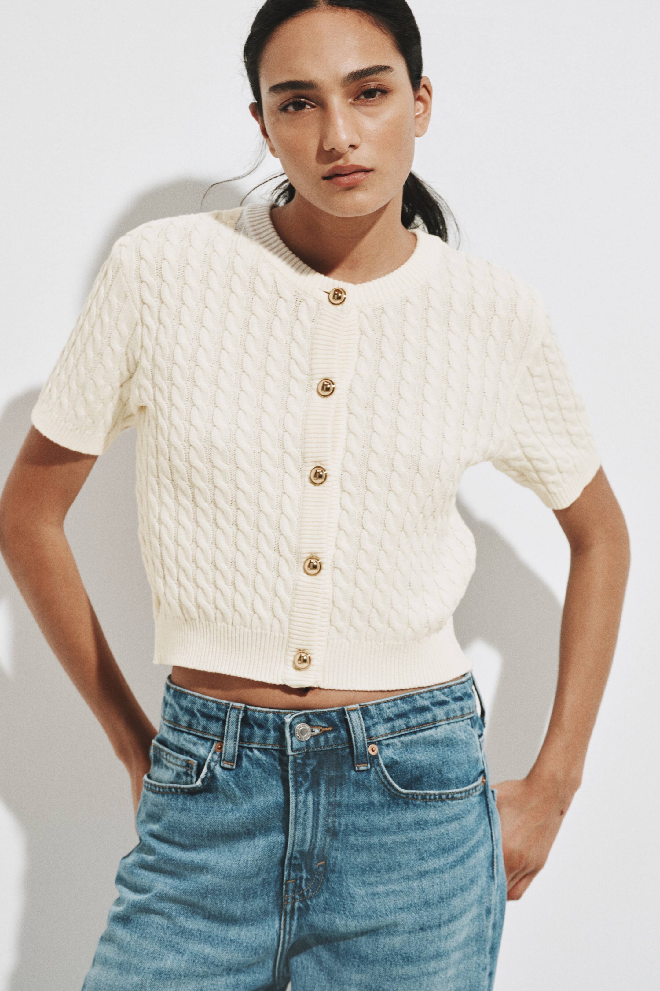 View larger image: A cream cable-knit cardigan features an all-over cable knit pattern, short sleeves and a round neckline. It presents a cropped silhouette with a ribbed hem, and gold-toned buttons adorn the front placket.