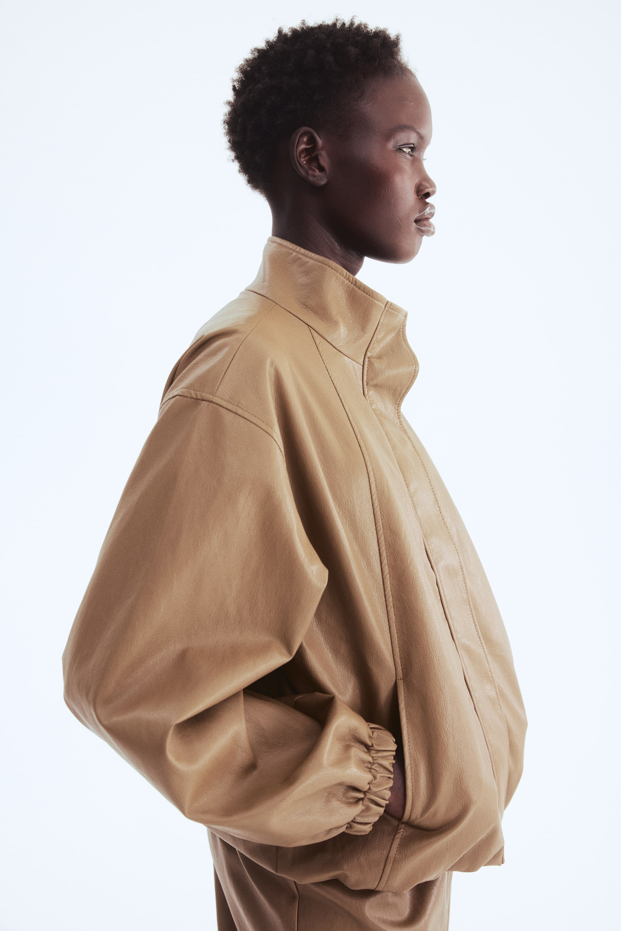 View larger image: The model is wearing an oversized jacket in tan coated fabric, casually slipping one hand into a discreet side pocket. The jacket showcases a stand collar, dropped shoulders, long sleeves with elasticated cuffs, and an elasticated hem that creates a puffy silhouette.