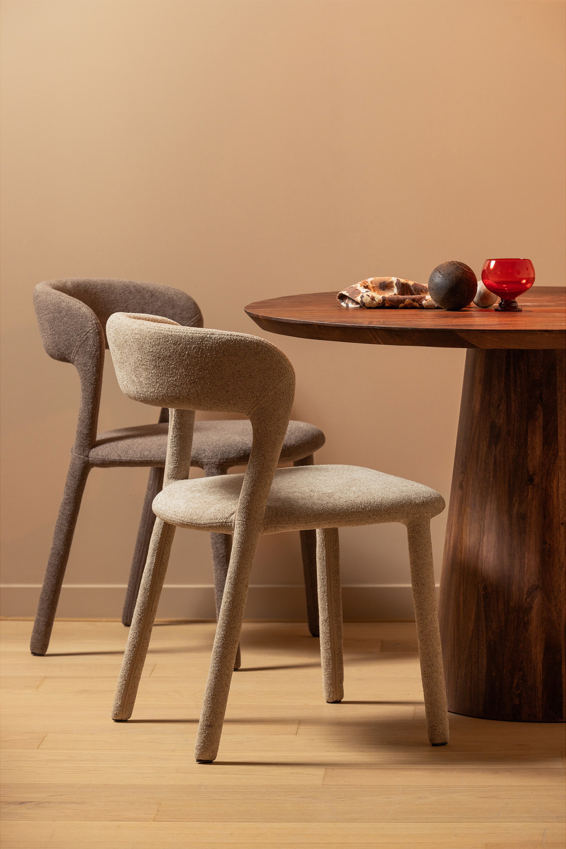 Zobacz większy obraz: Dwa tapicerowane krzesła tworzą zakrzywione oparcia i siedziska, z tapicerowanymi nogami płynnie przechodzącymi z głównej konstrukcji. Oba krzesła prezentują tkaninę o teksturze, przy czym jasnobeżowe krzesło jest lekko skierowane w lewo na pierwszym planie, a ciemnotaupe krzesło stoi za nim.