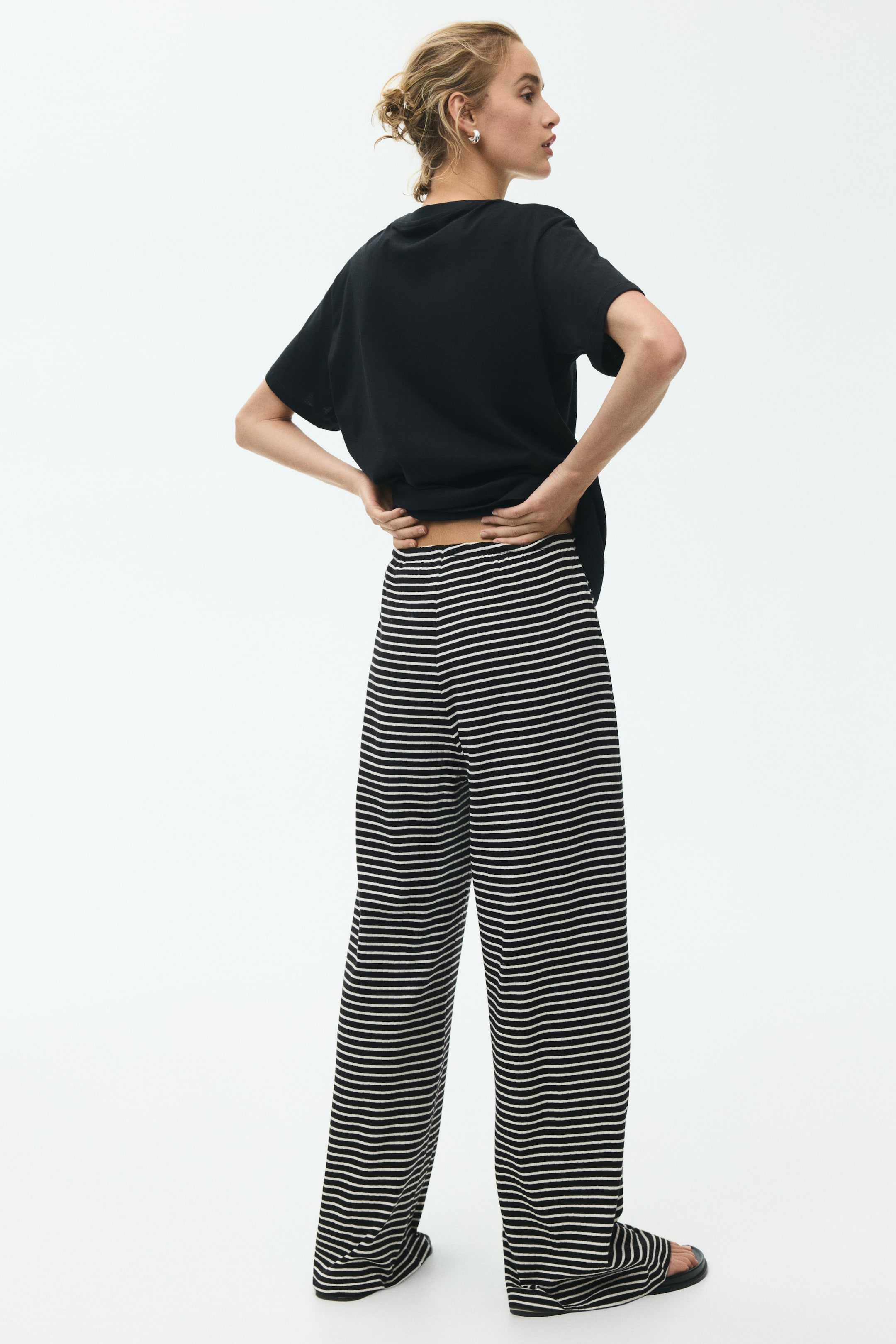 Ver imagen más grande: El modelo lleva unos pantalones de corte holgado en jersey de algodón acanalado a rayas blancas y negras. El modelo está ajustando la cintura elástica de los pantalones, que es visible al levantarse la camiseta negra.