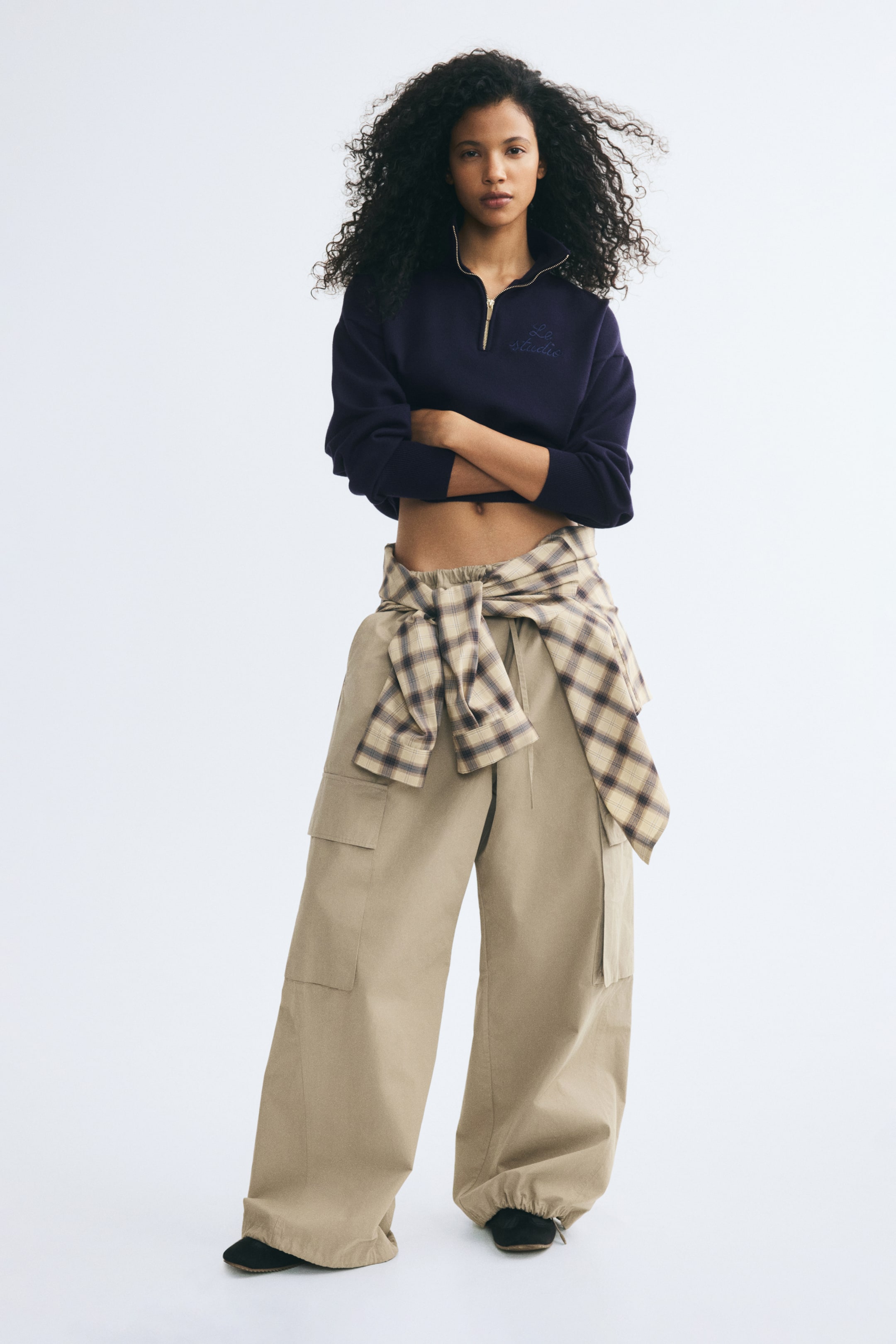 Visualizza immagine più grande: Un maglione a maglia blu navy dalla vestibilità ampia, con collo alto e mezza zip, spalle scese e dettaglio ricamato sul petto, indossato dalla modella. Il maglione corto è abbinato a pantaloni cargo beige a gamba ampia e a una camicia a quadri annodata in vita.