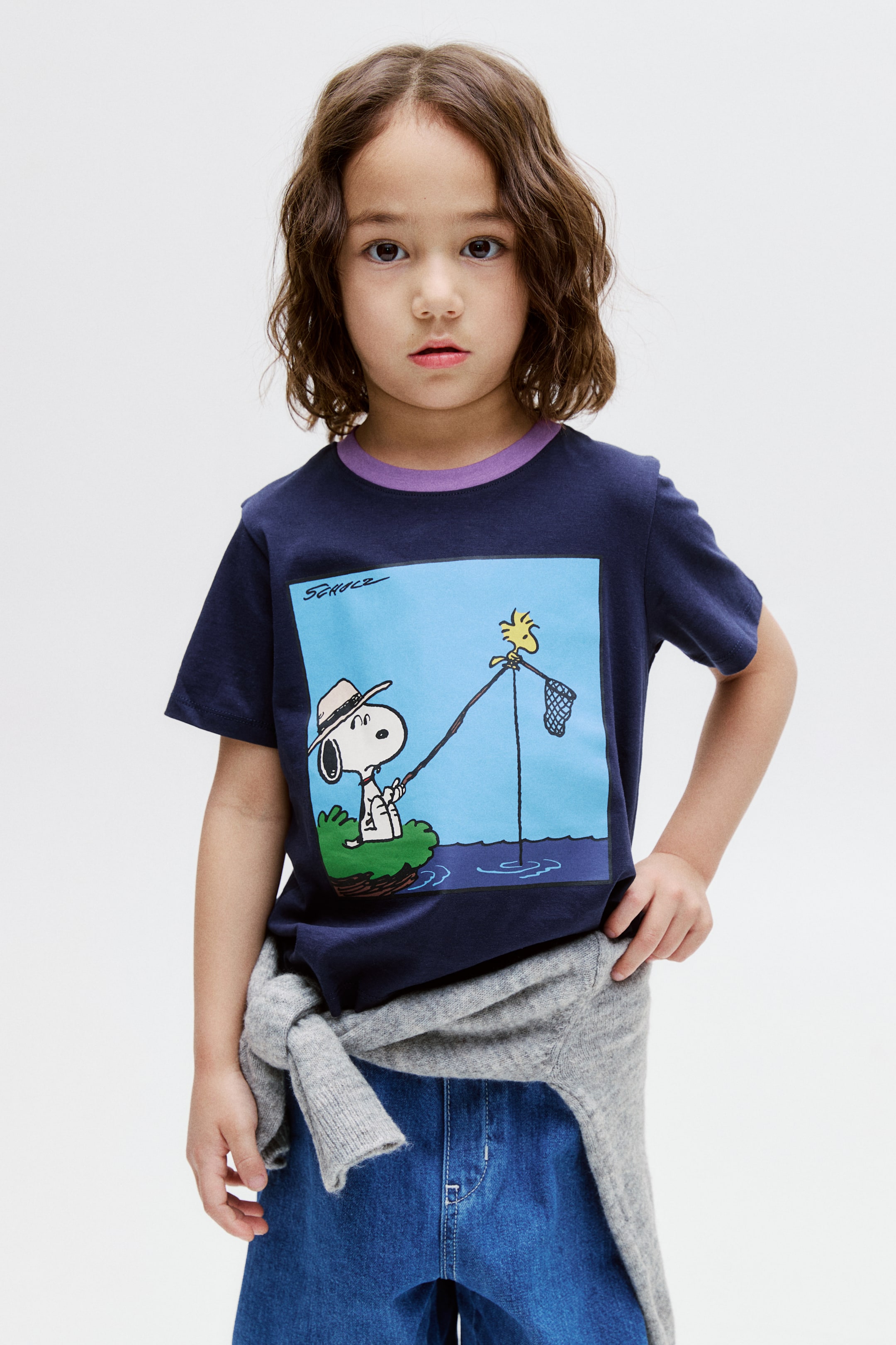 View larger image: "T-shirt biru navy ini menampilkan kolar berusuk ungu dan lengan pendek. Grafik berbentuk segi empat di bahagian depan menunjukkan Snoopy sedang memancing dari sepetak rumput, memakai topi, dengan Woodstock bertenggek di batang pancing sambil memegang jaring, dan tandatangan "SCHULZ" kelihatan di bahagian kiri atas."