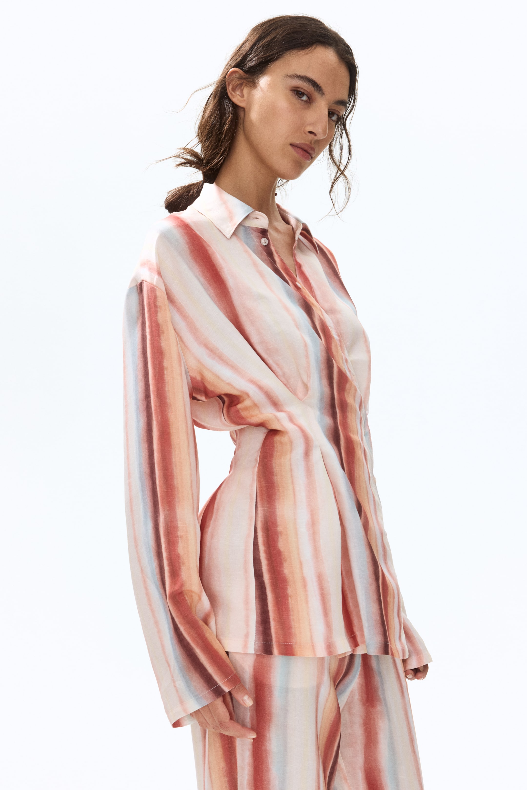 View larger image: The model is donning a shirt adorned with vertical watercolor-esque stripes in hues of pink, orange, brown, and light blue. This shirt features a collar, dropped shoulders, and long sleeves. The wrapover front showcases an angled button band, while the box pleats at the waist create a cinched-in effect.