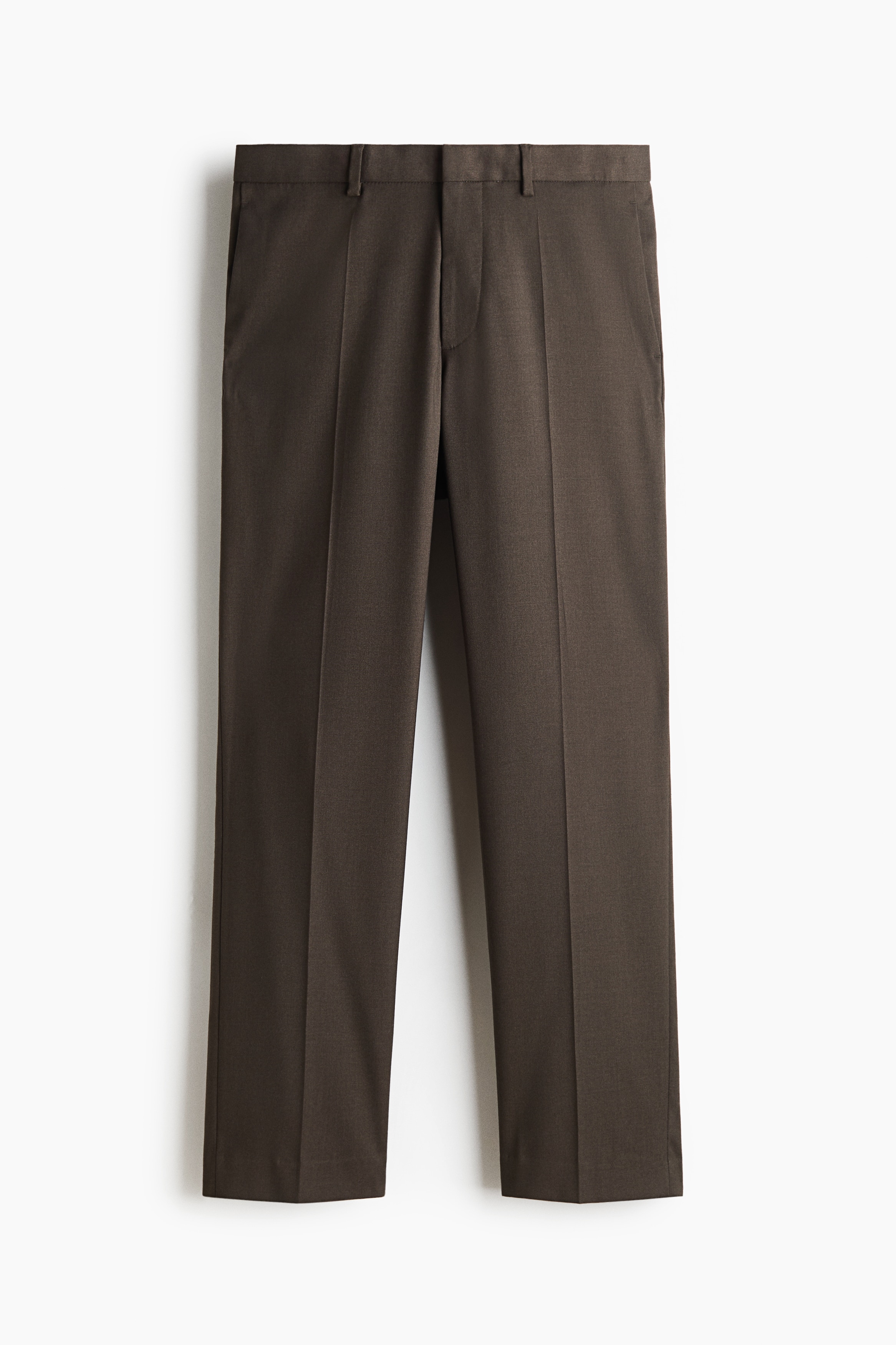 A dark brown suit, comprising a single-breasted blazer and matching tailored trousers, is worn by the model with hands placed in the trouser pockets. The suit is layered over a black crewneck top and paired with black leather loafers.