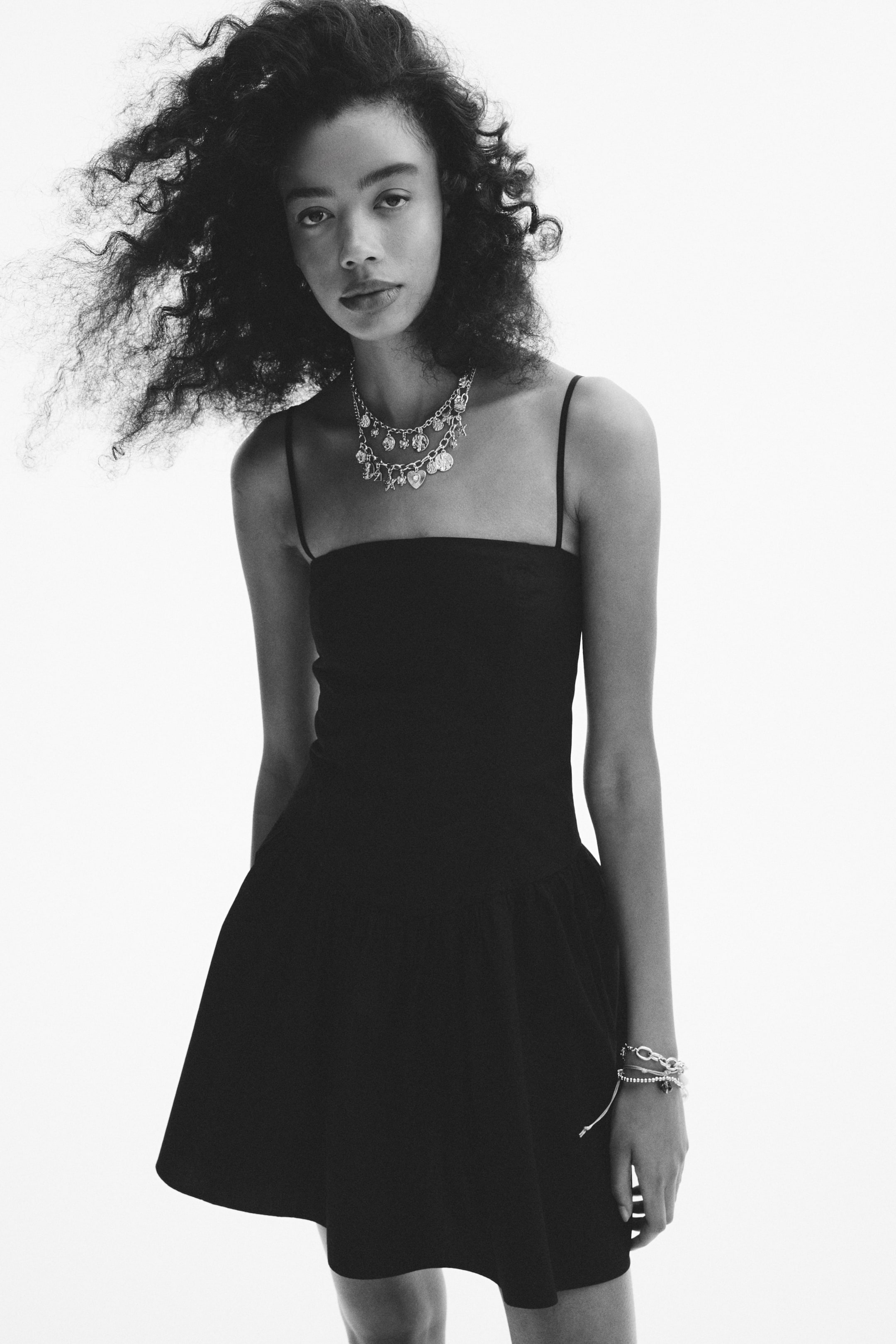 View larger image: A black mini-dress na may manipis na spaghetti straps, fitted na bodice, at flared na palda. Isang layered na silver-toned charm necklace ang nakasuot sa leeg, at maraming silver-toned bracelets ang nasa kaliwang pulso.
