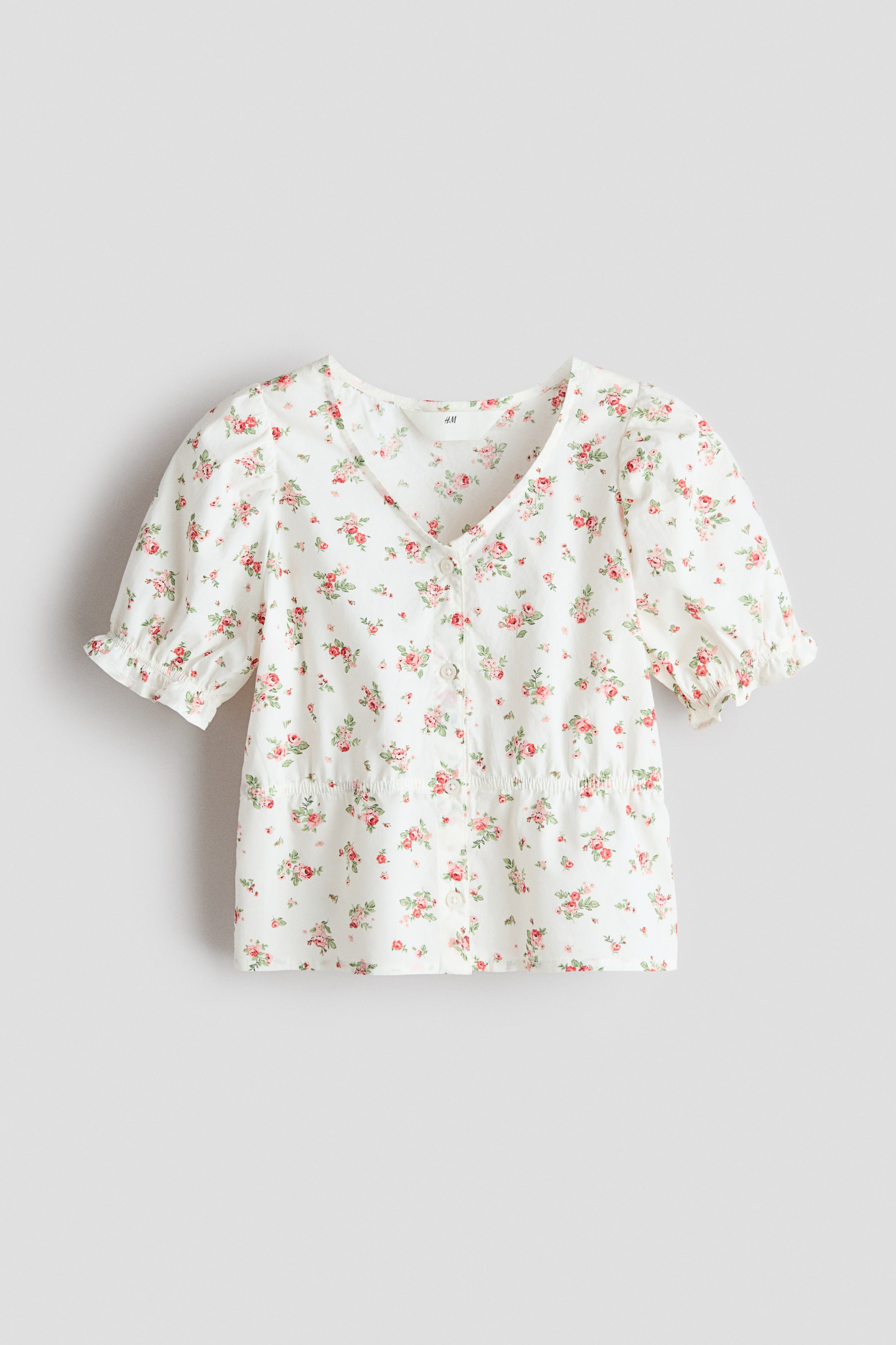 View larger image: A white blouse crafted from airy cotton seersucker showcases its front, adorned with a floral motif of red and pink roses, a V-shaped neckline, and buttons running down the front. It features narrow elastication at the waist and short puff sleeves with elasticated, frill-trimmed cuffs.