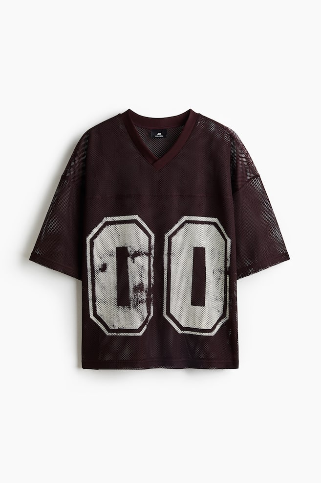 Un t-shirt en maille brun foncé présente une coupe ample et surdimensionnée avec un col en V, des épaules tombantes et un ourlet droit. Le DEVANT affiche de grands chiffres blancs usés '00' sous un empiècement visible. - 1