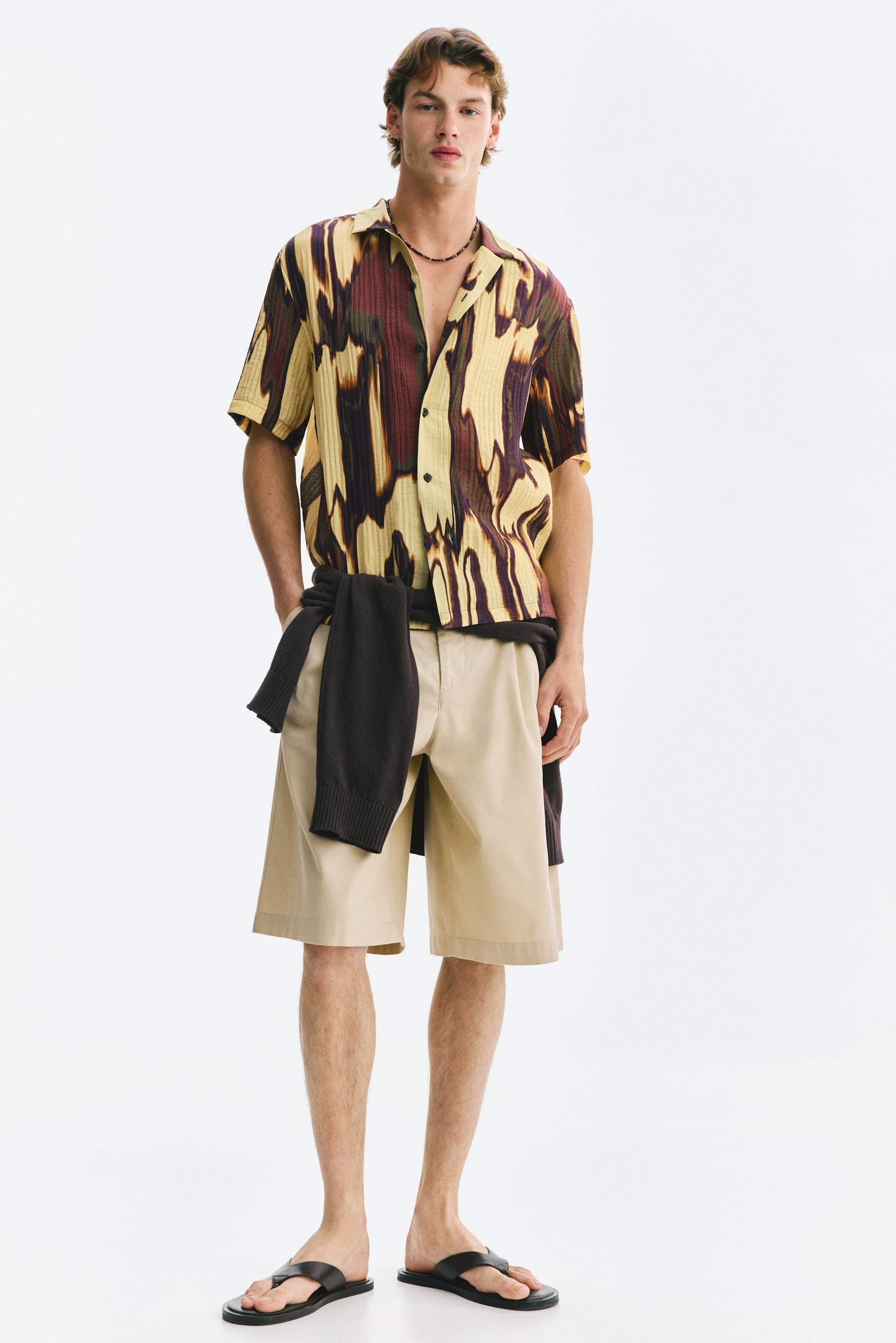 View larger image: A short-sleeved, collared shirt featuring a textured finish with an abstract pattern in shades of yellow, burgundy, and dark brown is paired with light beige, knee-length shorts. A dark brown knit sweater is casually tied around the waist, while black leather flip-flops complete the look.