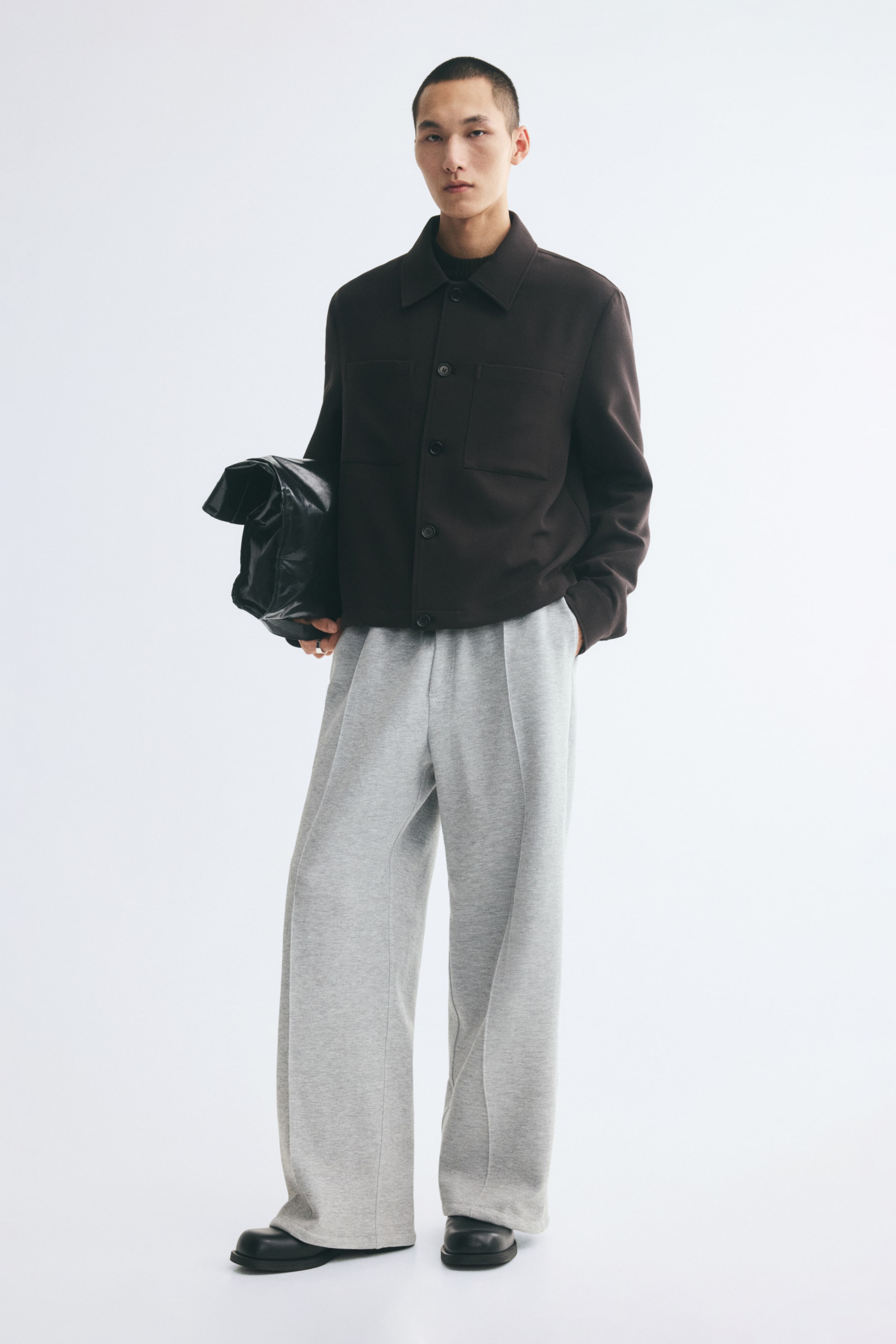 View larger image: Grey track pants with a relaxed fit, straight legs, and stitched front creases are worn by the model, who is standing with one hand in a side pocket. The pants are styled with a dark brown buttoned jacket, black leather shoes, and a black clutch bag tucked under one arm.