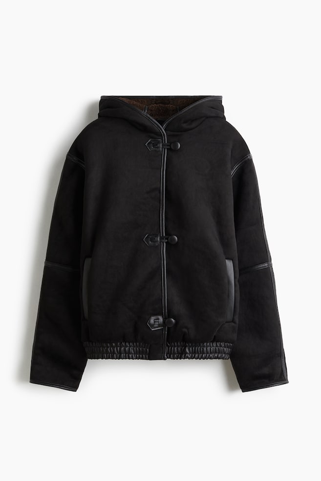 Eine schwarze Kapuzenjacke wird von vorne präsentiert, mit einer matten Außenseite und schwarzer Paspelierung entlang der Kanten, Nähte und um drei zentrale Knebelverschlüsse. Sie verfügt über seitliche Schlitztaschen und einen gerippten Saum, wobei die Kapuze ein braunes Kunstfellfutter zeigt. - 1