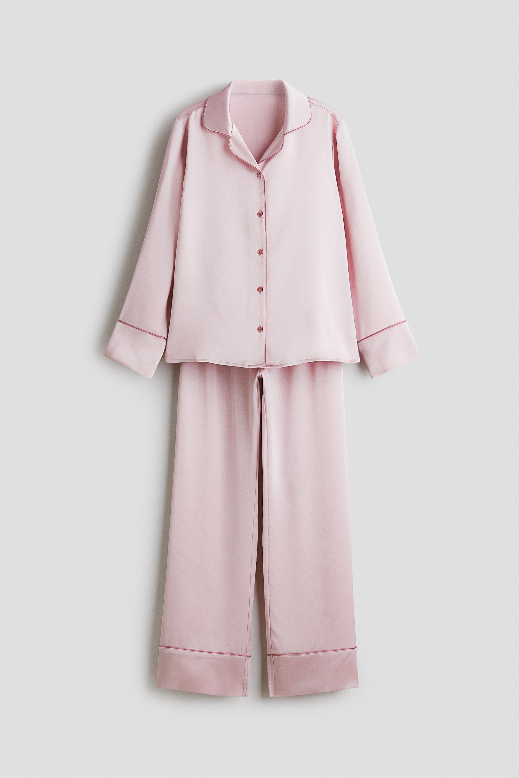 Größeres Bild ansehen: Ein rosa Satin-Pyjama-Set mit einem langärmeligen Oberteil mit Knopfleiste vorne, Reverskragen, passenden rosa Knöpfen und dunkelrosa Paspeln entlang des Kragens, der Knopfleiste und der Manschetten. Die dazu passenden langen Hosen zeigen ebenfalls dunkelrosa Paspeln an den Säumen, wobei das gesamte Set von vorne präsentiert wird.