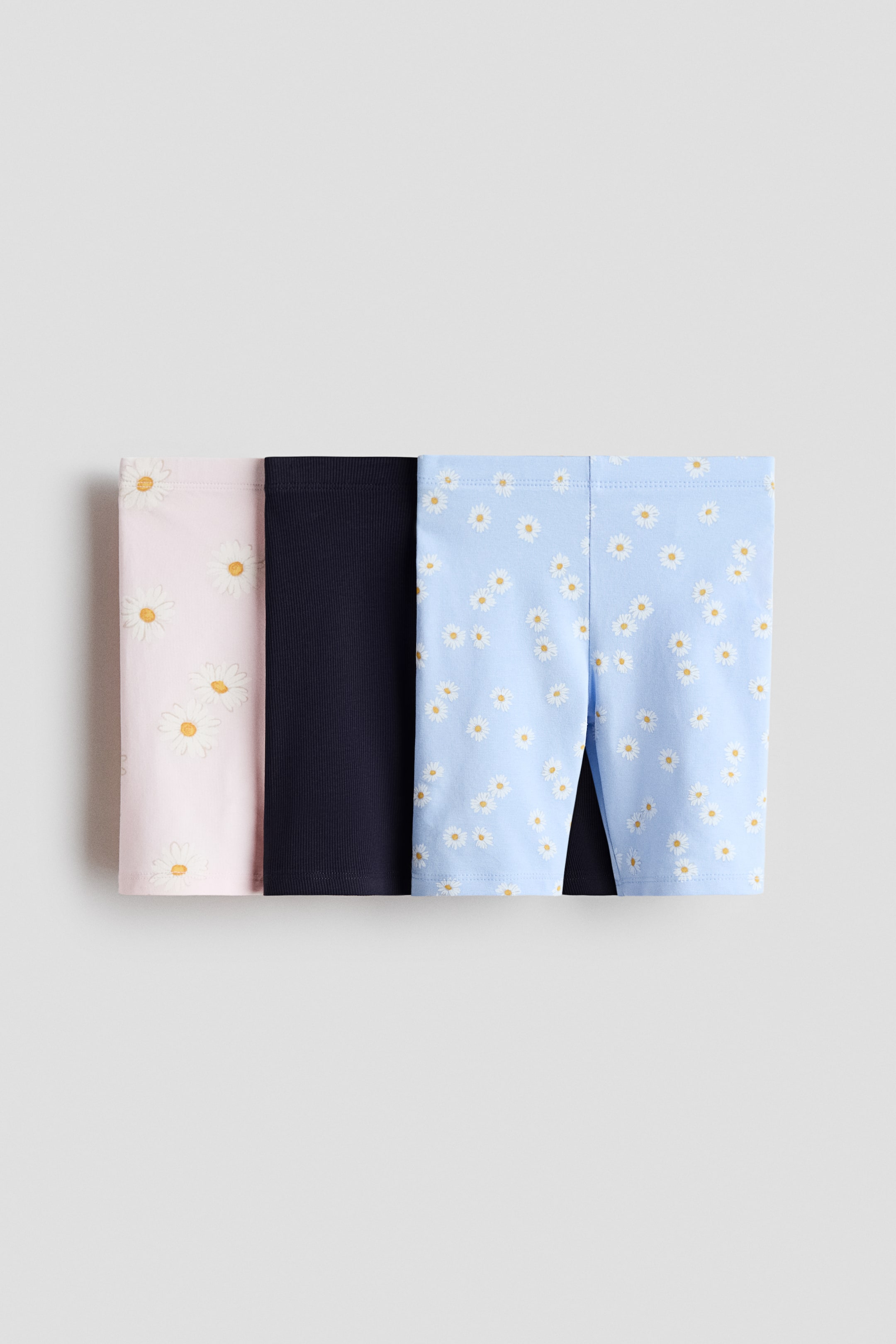 View larger image: Three pairs of shorts are showcased, displaying their front. The first pair is in a light pink hue adorned with a white daisy motif, the second pair is a solid navy blue featuring a ribbed texture, and the third is light blue with a white daisy motif.