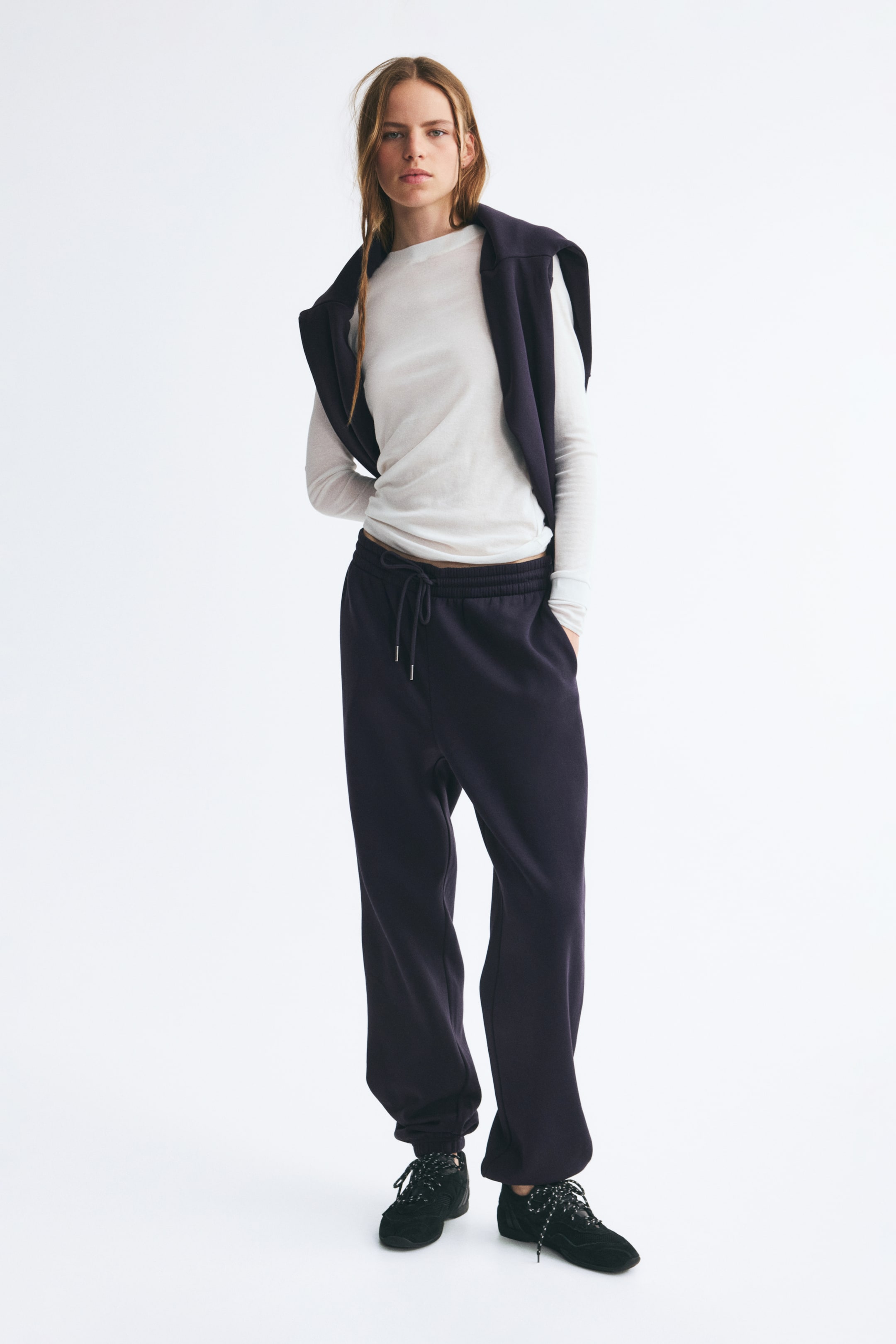 View larger image: Dark purple track pants featuring a high, elasticated waistband with a drawstring and gathered cuffs are worn by the model. The model is standing with one hand resting in the side pocket, styling the pants with a white long-sleeve shirt, a matching dark sweater draped over the shoulders, and black sneakers.