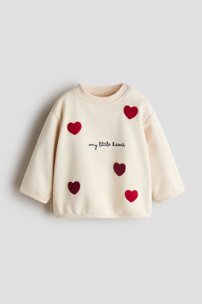 Sweter Design Detail jest ustawiony przodem, prezentujÄc kremowÄ tkaninÄ z prÄÅ¼kowanym okrÄgÅym dekoltem, prÄÅ¼kowanymi mankietami i prÄÅ¼kowanym doÅem. PiÄÄ czerwonych fakturowanych aplikacji w ksztaÅcie serca otacza centralny czarny haftowany napis âmy little heartâ. - 1