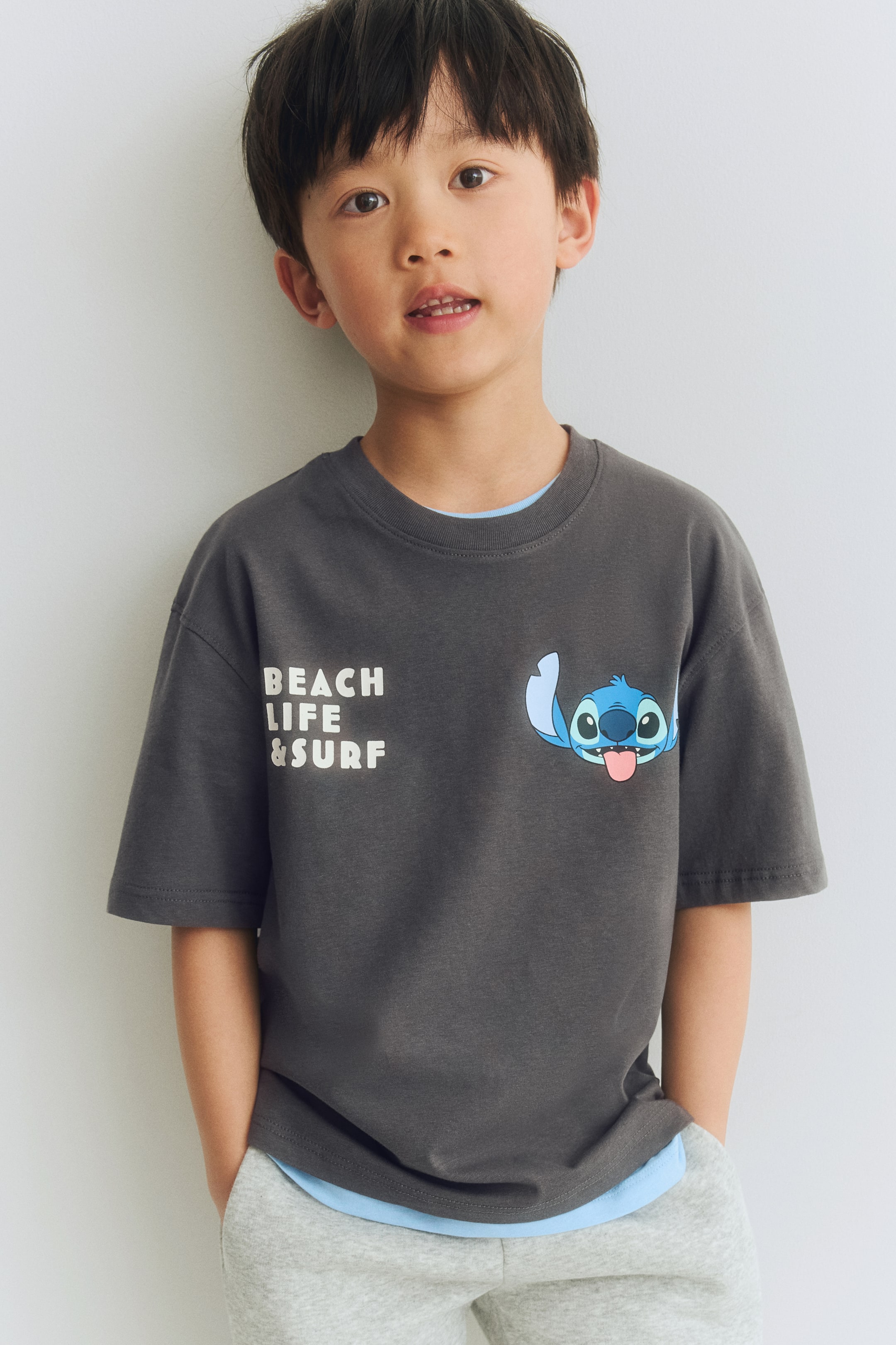 View larger image: A dark grey t-shirt featuring a round neck and short sleeves, displaying white text reading 'BEACH LIFE & SURF' on the left chest and a blue cartoon character with its tongue out on the right chest. A light blue t-shirt is layered underneath, visible at the neckline and hem of the dark grey t-shirt.