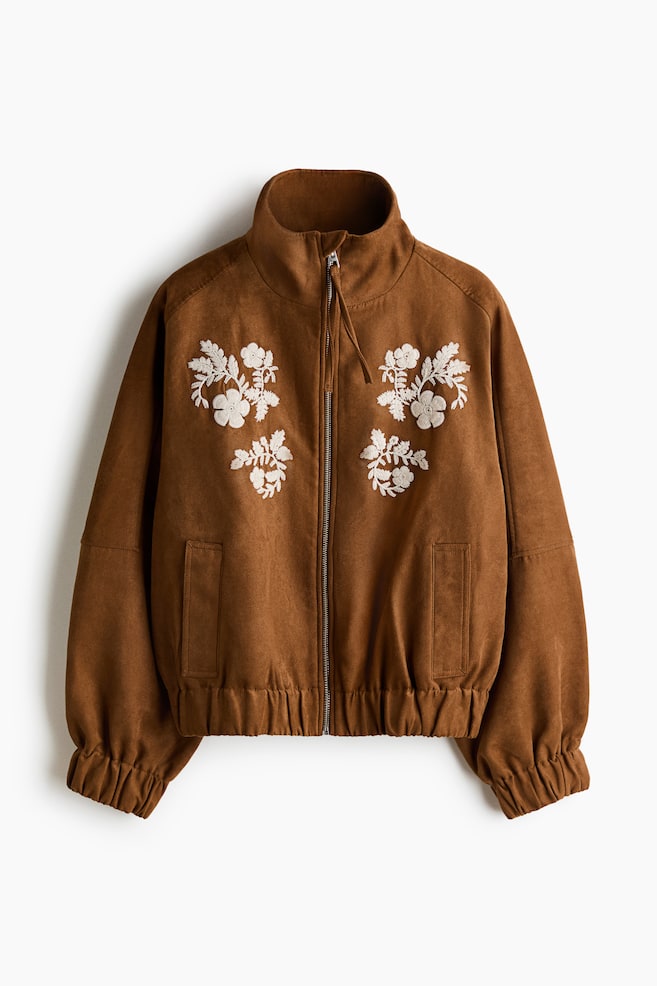 A brown suede-like jacket is shown from the front, featuring white floral embroidery on both sides of the chest. The jacket has a high stand-up collar, a front zipper closure with a pull tab, two vertical welt pockets, and gathered elastic detailing at the hem and cuffs. - 1