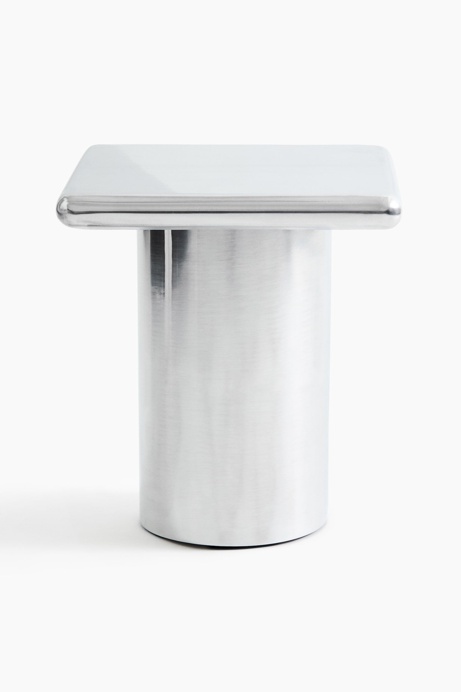 A metallic side table is shown in a front-facing view, featuring a square tabletop with rounded corners and a thick, cylindrical pedestal base. The table has a brushed silver finish with a semi-polished, reflective top surface. - 1