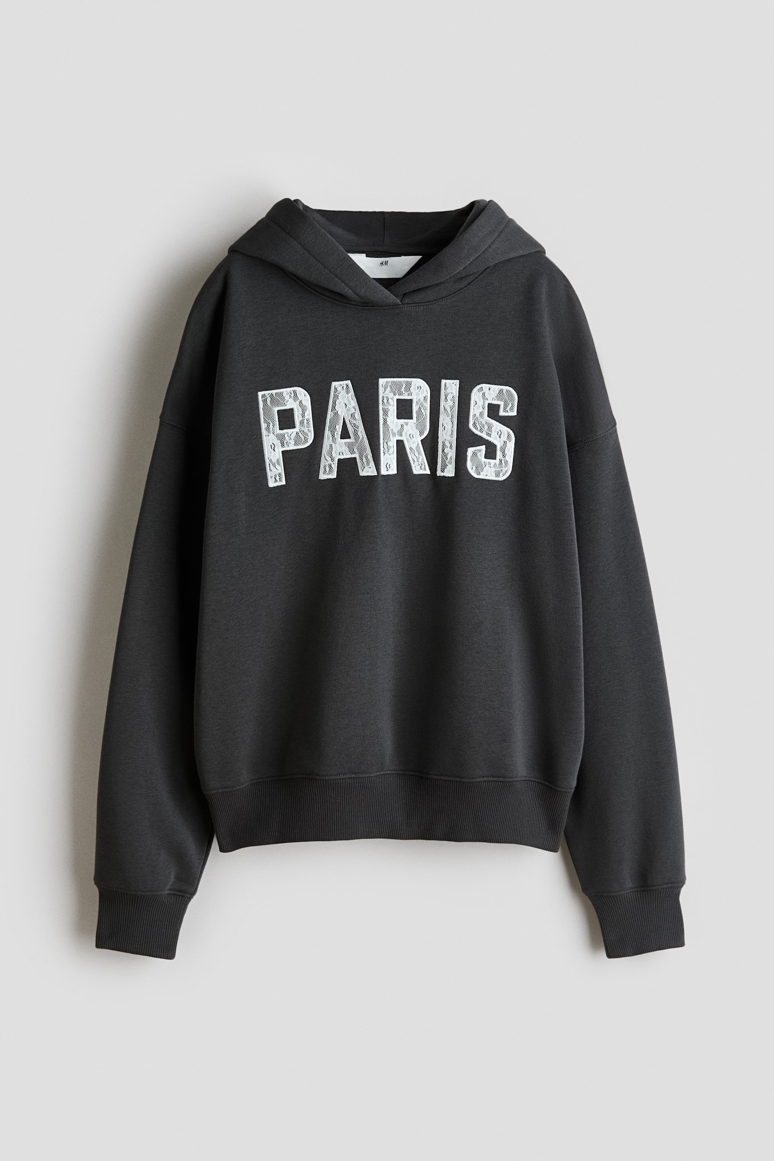Een donkergrijze hoodie wordt van voren getoond en laat het woord "PARIS" zien in witte blokletters met een kanten inzetstuk over de borst. Het kledingstuk heeft een wikkelkap, verlaagde schouders, lange mouwen met ribgebreide boorden en een ribgebreide zoom.