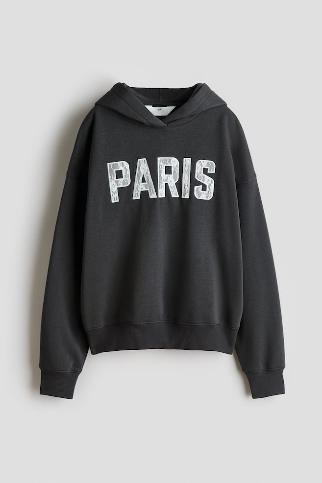 Ein dunkelgrauer Hoodie wird von vorne gezeigt und prÃ¤sentiert das Wort âPARISâ in weiÃen Blockbuchstaben mit einer Spitzenapplikation quer Ã¼ber der Brust. Das KleidungsstÃ¼ck verfÃ¼gt Ã¼ber eine Wickelkapuze, Ã¼berschnittene Schultern, lange Ãrmel mit RippbÃ¼ndchen und einen gerippten Saum. - 1