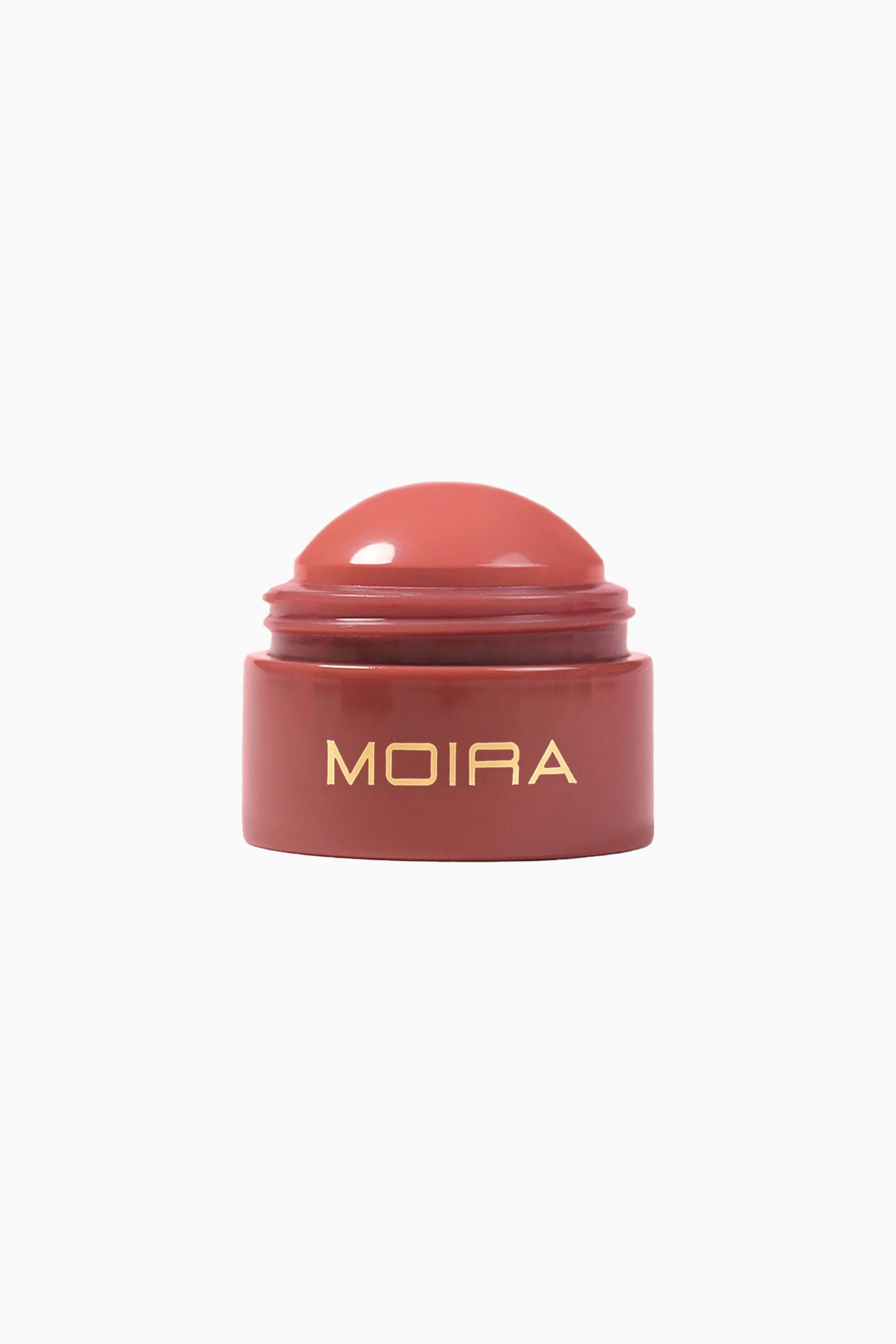 Näytä suurempi kuva: Moira Soft Blush Balm on asetettu pystyyn eteenpäin suuntautuvaan näkymään, jossa näkyy kupolimainen, lämpimän ruusunvärinen voidemainen balmi, joka nousee esiin sitä vastaavasta sylinterimäisestä pakkauksesta. Pakkauksen etupuolella näkyy tuotemerkin nimi ”MOIRA” kullanvärisellä tekstillä.