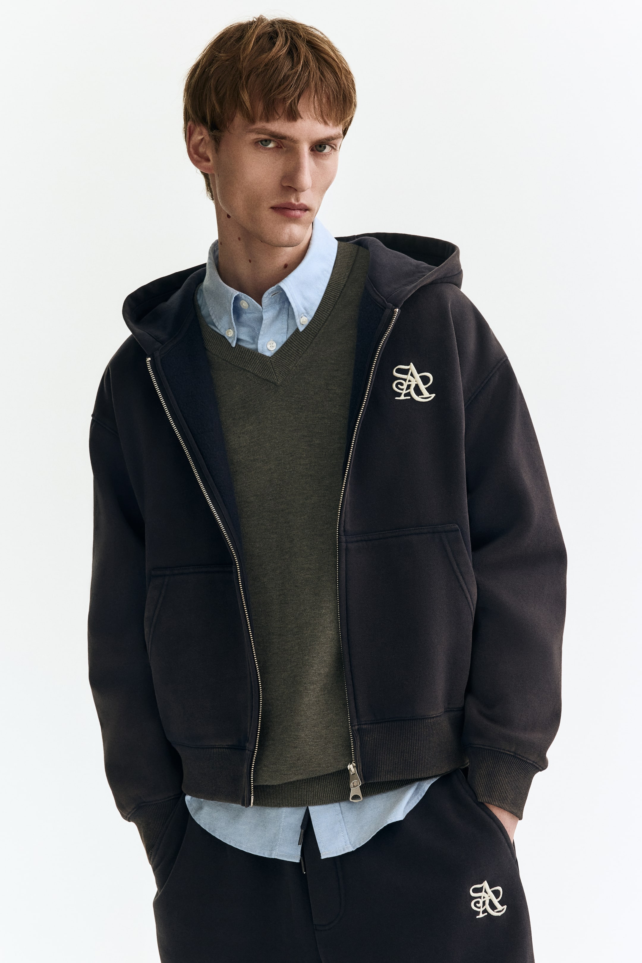 Visa större bild: En mörk huvtröja med dragkedja bärs, med en huva, en metallblixtlås som löper längs framsidan, ribbade muddar och fåll samt integrerade sidofickor. Ett broderat logotyp syns på vänster bröst, och huvtröjan är stylad öppen över en V-ringad tröja och en skjorta med krage.