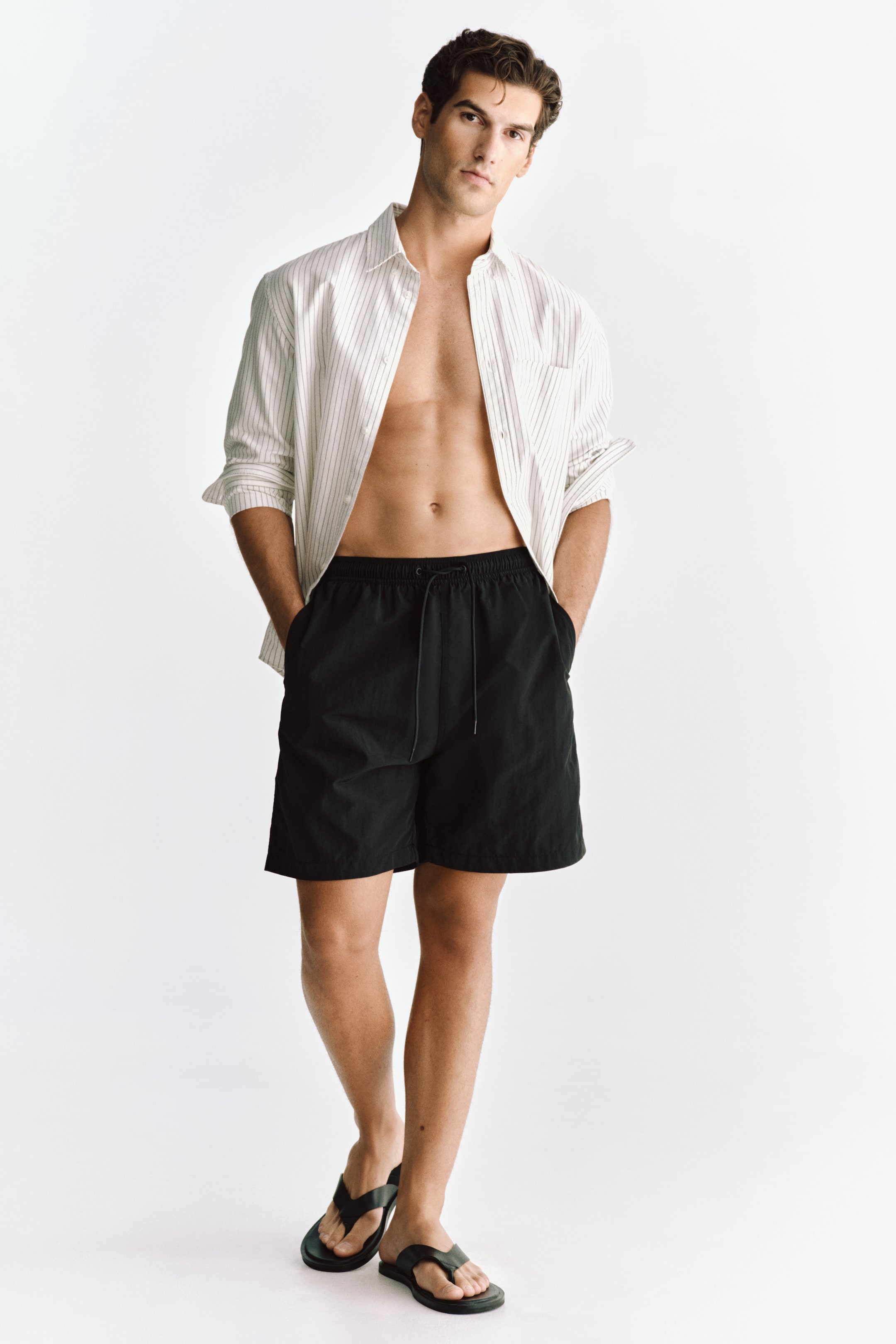 Voir une image plus grande: Le modèle porte un short de bain en nylon noir, doté d'une taille à cordon de serrage avec élastication recouverte et de poches latérales. Les mains du modèle reposent dans les poches latérales du short, qui se termine au-dessus des genoux.
