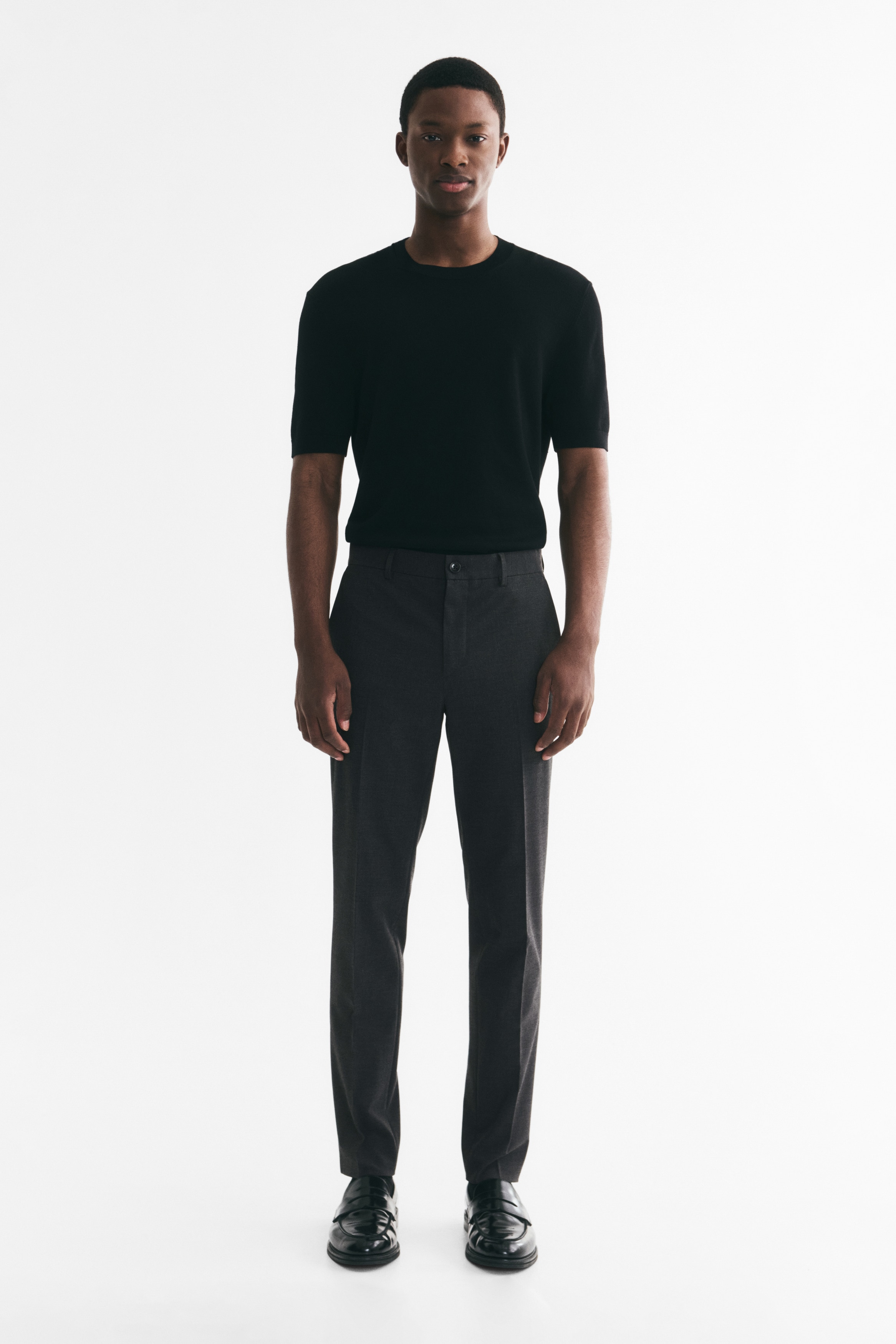 Dark gray tailored trousers in a soft weave are presenting a front view. They are featuring a zip fly with a single button closure, side pockets, and sharp creases running down the front of each leg, creating a slim, fitted silhouette.