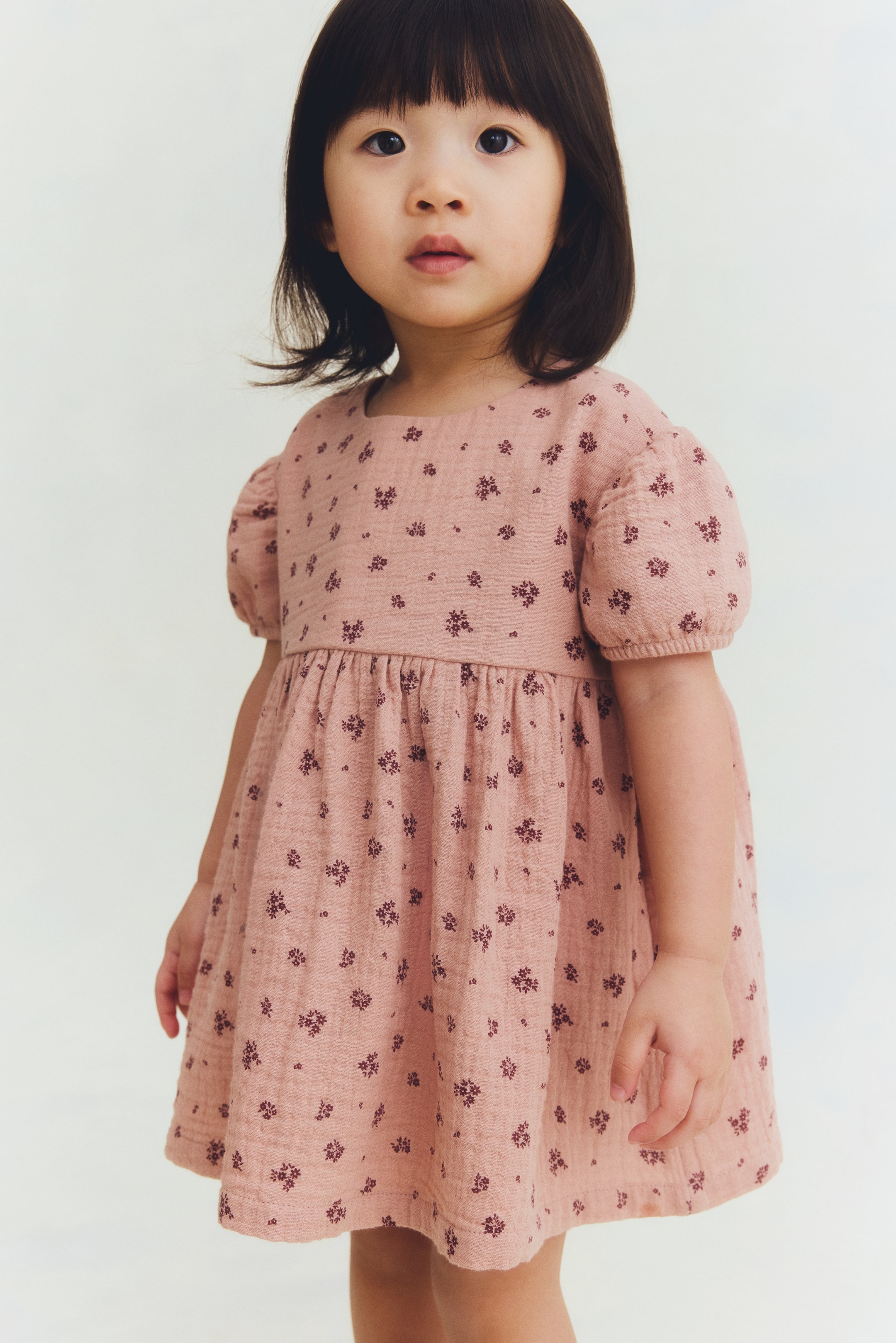 View larger image: A child's dusty rose dress showcases a textured fabric adorned with a subtle dark floral print. It is crafted with a round neckline, puffed short sleeves, and a gathered waistline, cascading into a flared skirt.
