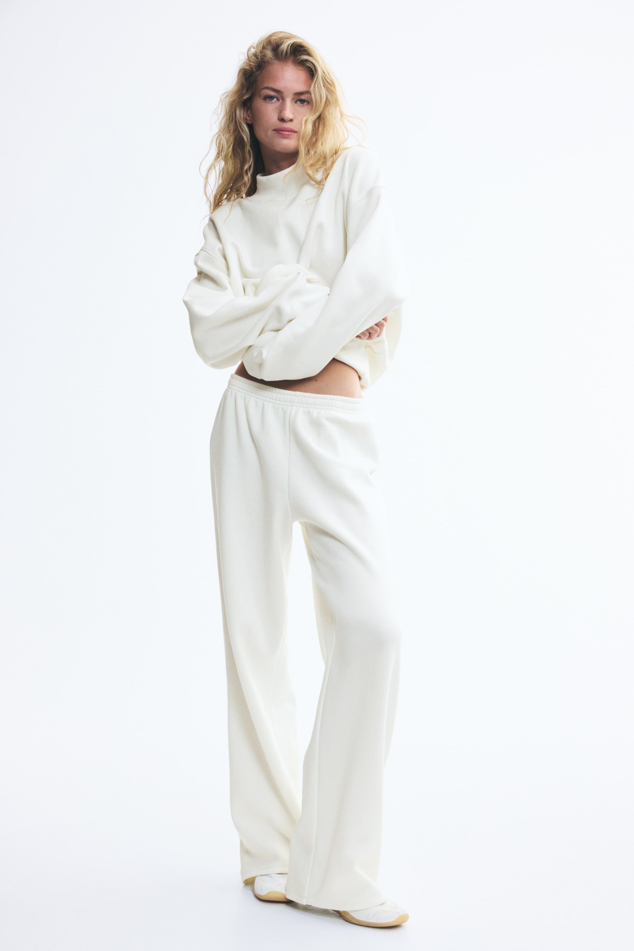Visualizza l'immagine ingrandita: Il modello indossa un set lounge coordinato color bianco sporco, composto da un maglione con collo alto e pantaloni da tuta dritti. Il maglione presenta un collo alto a coste, spalle scese e polsini a coste, mentre i pantaloni da tuta sono dotati di vita elasticizzata e una vestibilità rilassata e dritta.