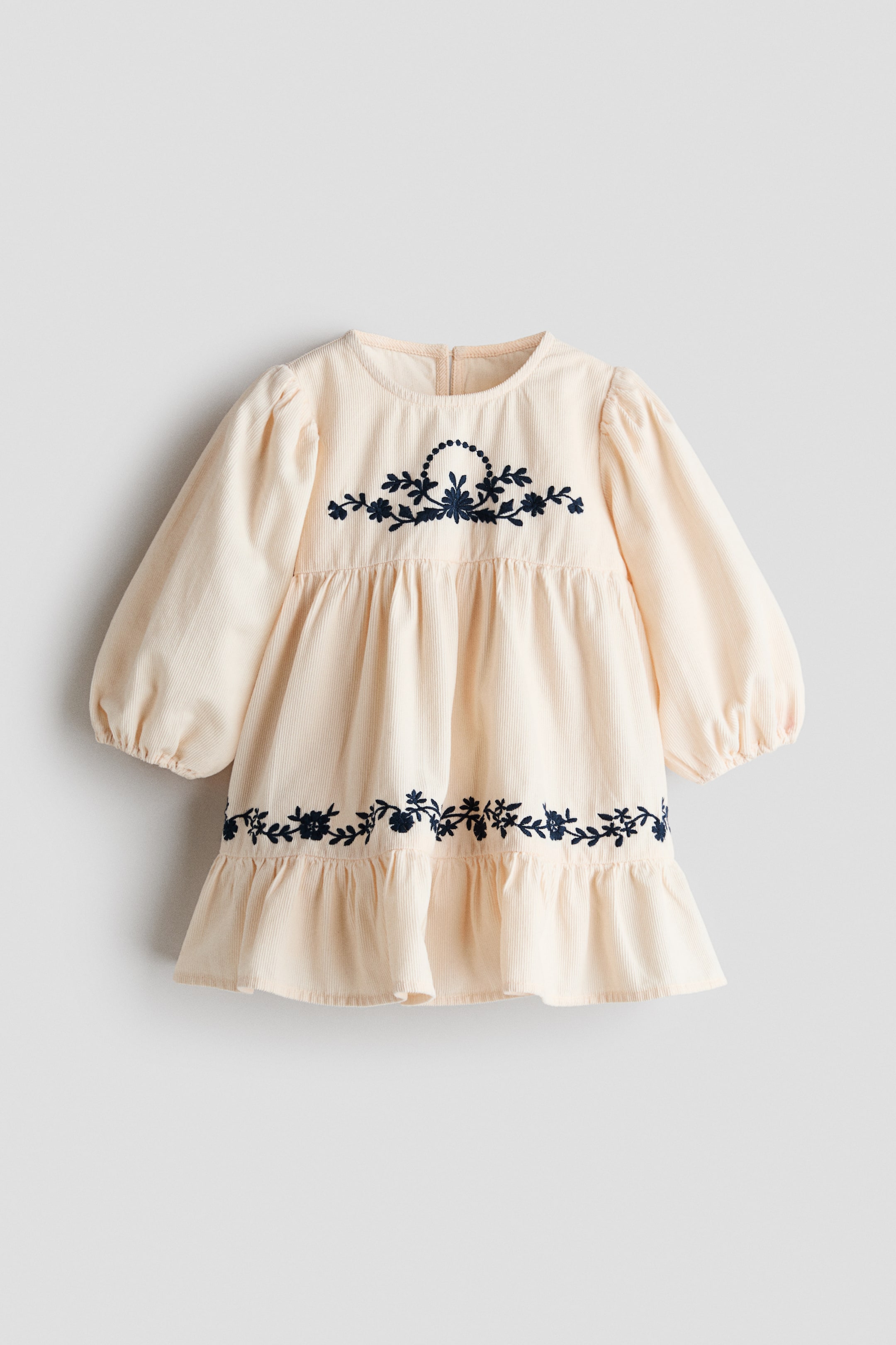 View larger image: A cream-coloured corduroy dress is laid flat, showcasing dark blue floral embroidery across the chest and just above the ruffled hem. The dress features a round neckline, long balloon sleeves with elastic cuffs, and a gathered empire waist.