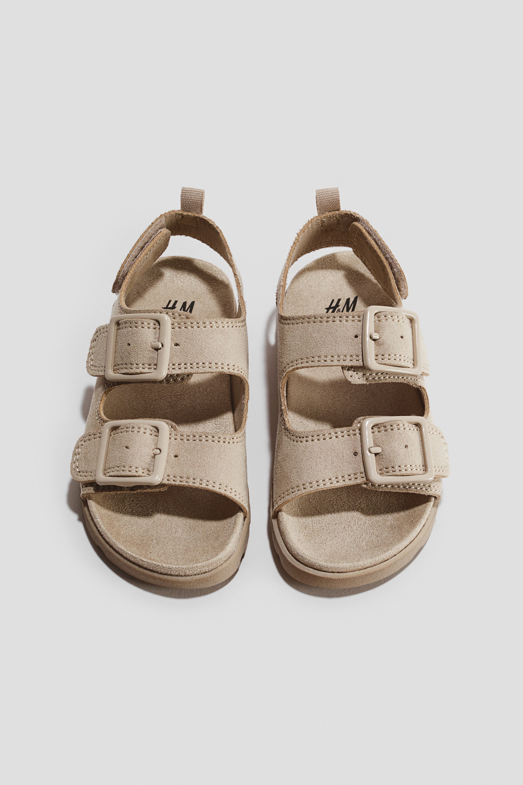 View larger image: A pair of beige, suede-textured sandals is shown from a top-down front angle, featuring two wide adjustable straps across the foot with matching plastic buckles. The sandals are designed with a moulded footbed and a slingback ankle strap with a small pull tab at the heel.