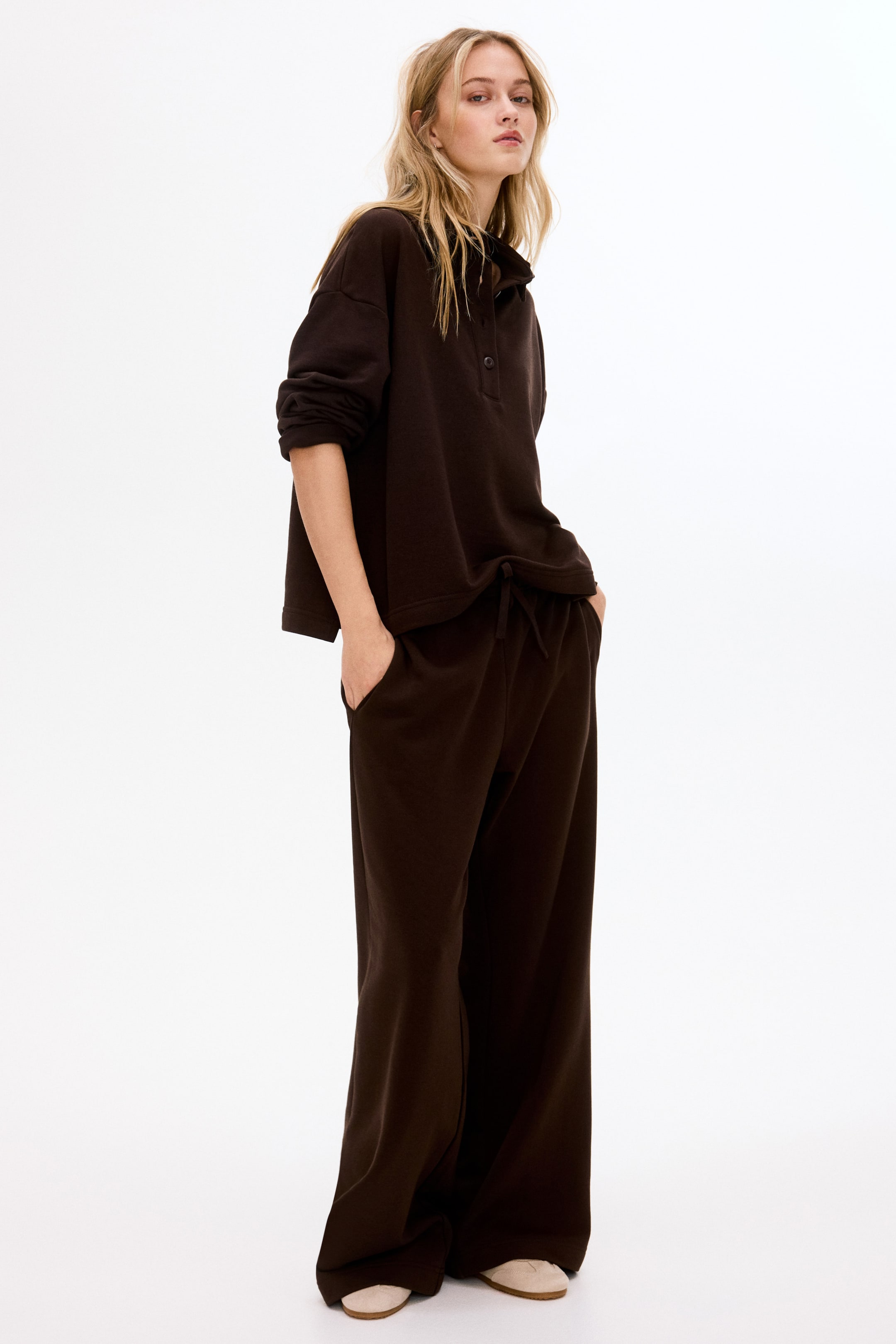 Grotere afbeelding bekijken: Een donkerbruin tweedelig loungewear-set wordt getoond, bestaande uit een longsleeve trui met een polokraag en knoopsluiting, en wijde broek met een trekkoord in de taille.