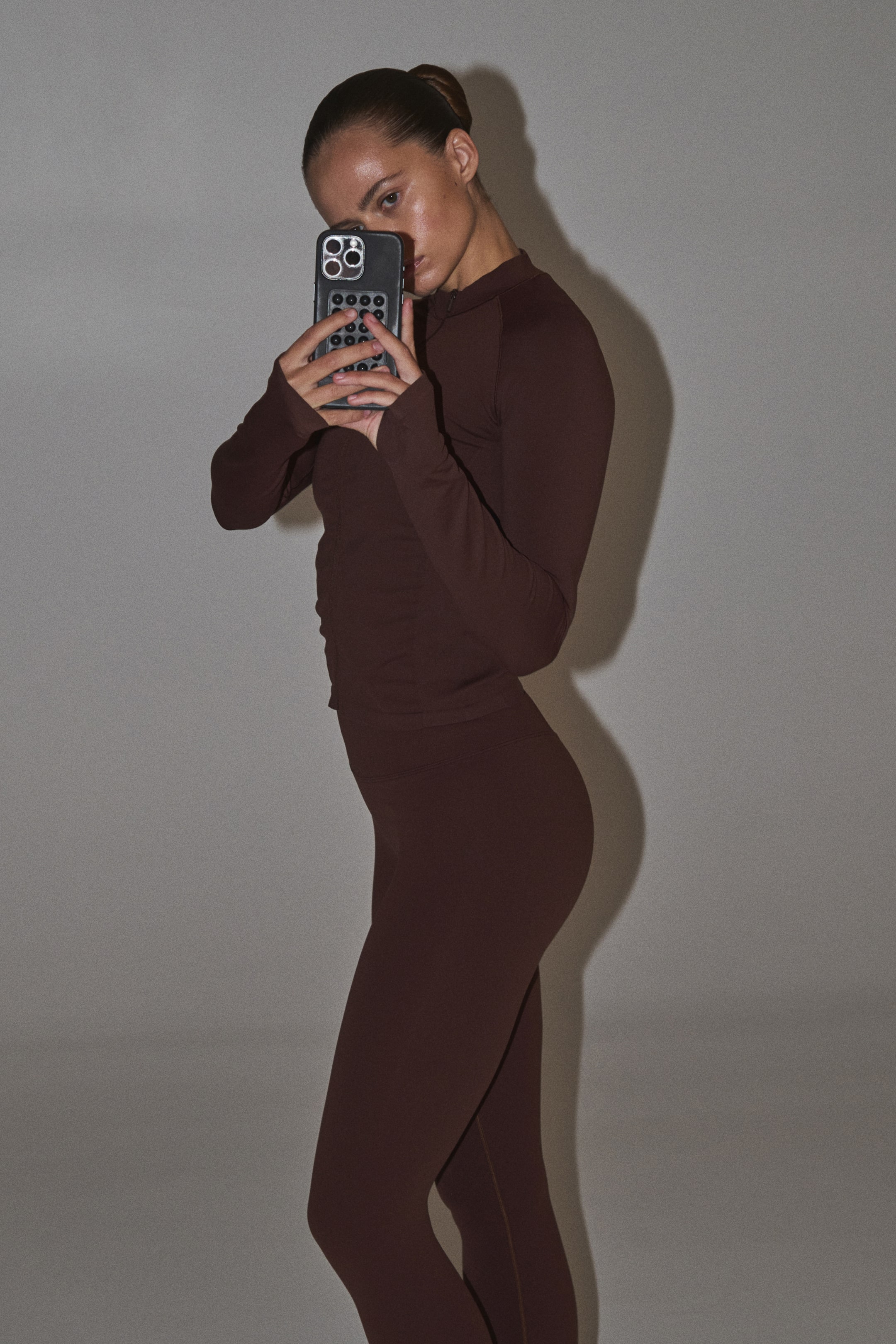 View larger image: A dark-brown long-sleeved top and matching high-waisted leggings are shown from the right side. The top features a high neckline and raglan sleeves, with both pieces fitting snugly to the body.