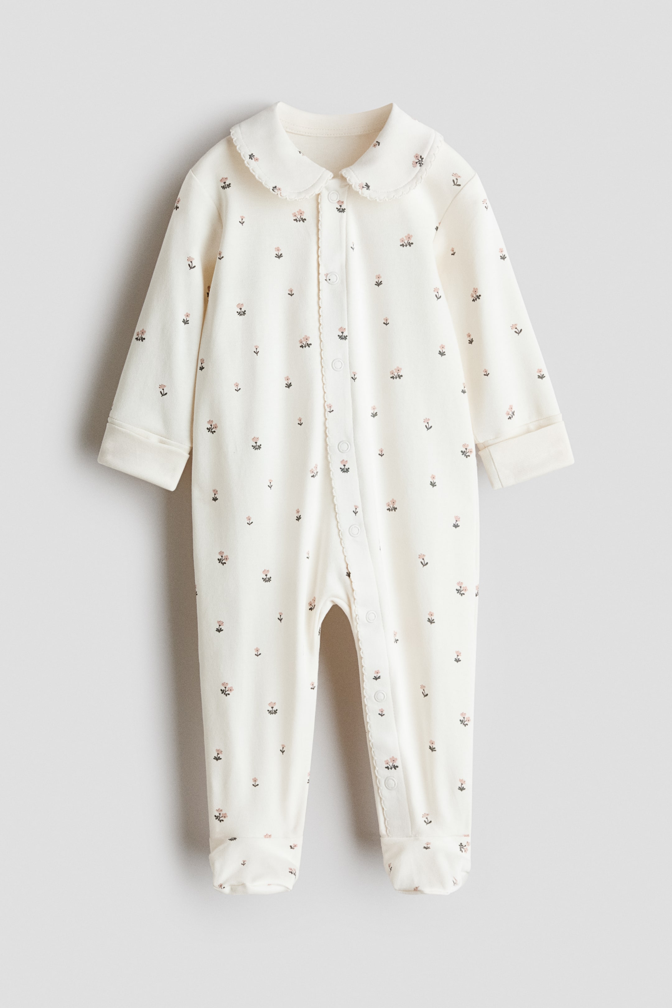 View larger image: An off-white baby sleepsuit is shown from the front, featuring a delicate all-over pink floral pattern. The design includes a Peter Pan collar with scalloped trim, a front snap-button closure with a scalloped placket extending down one leg, long sleeves with wide cuffs, and integrated footies.