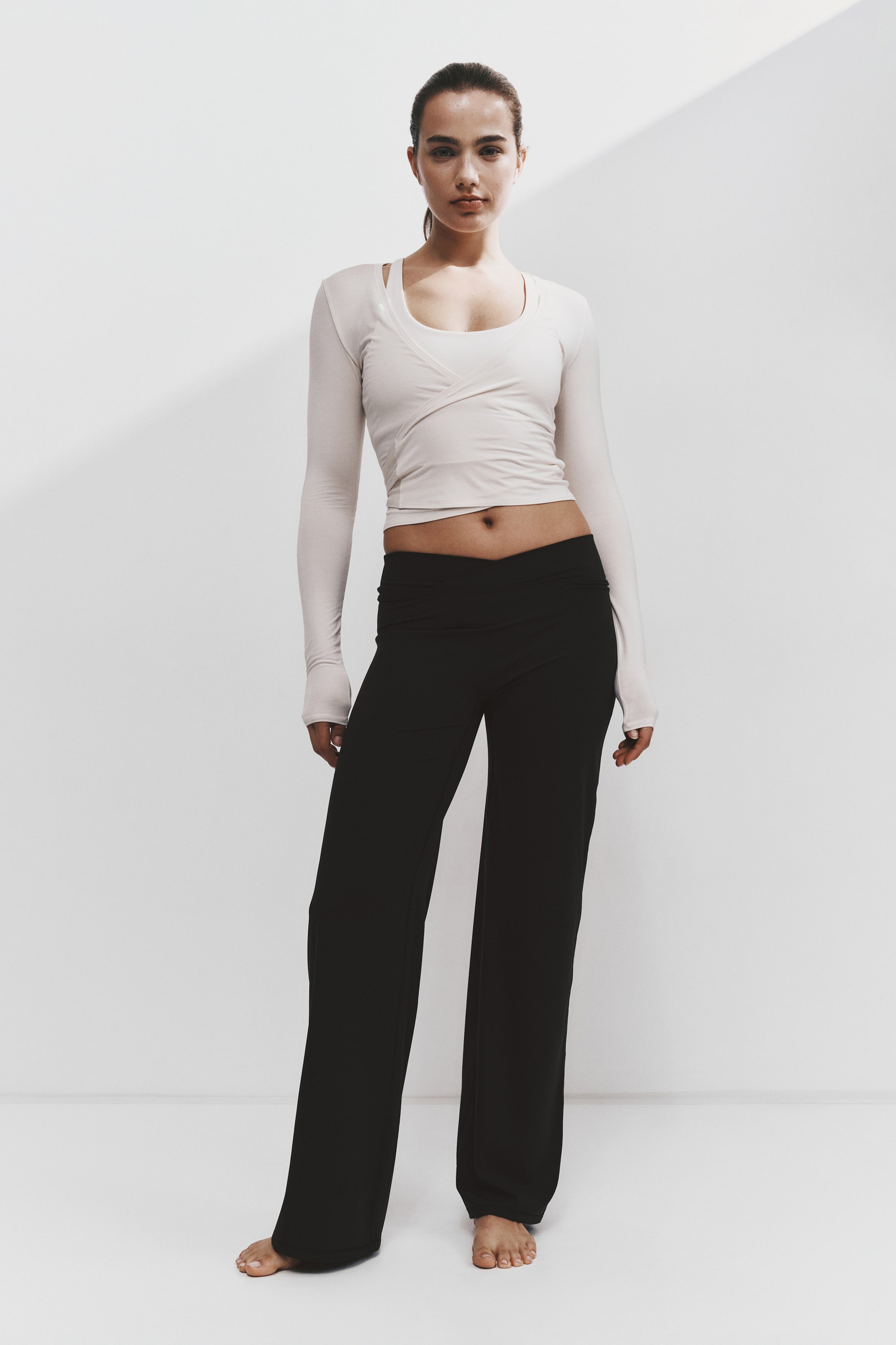 Agrandir l'image: Cette tenue de sport de face se compose d’un haut court beige clair à manches longues, doté d’un design portefeuille croisé et d’un col rond, associé à un pantalon noir à jambes larges avec une taille repliée.