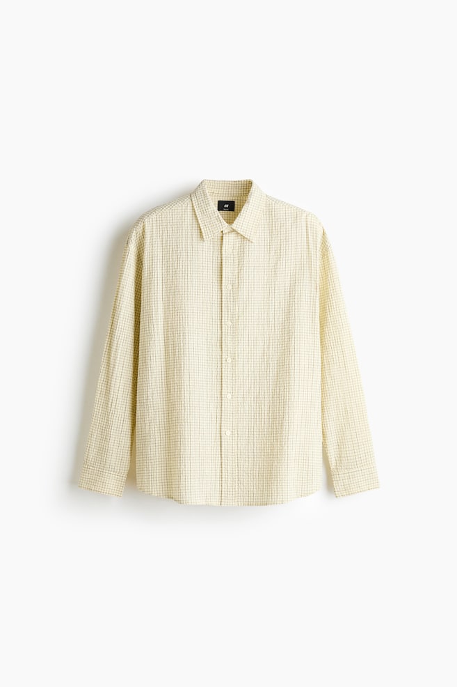A long-sleeved shirt in a textured, pale yellow checked weave is displayed in a relaxed fit from the front, featuring a turn-down collar and a classic button-up front. The shirt is designed with long sleeves, buttoned cuffs, and a gently rounded hem. - 1