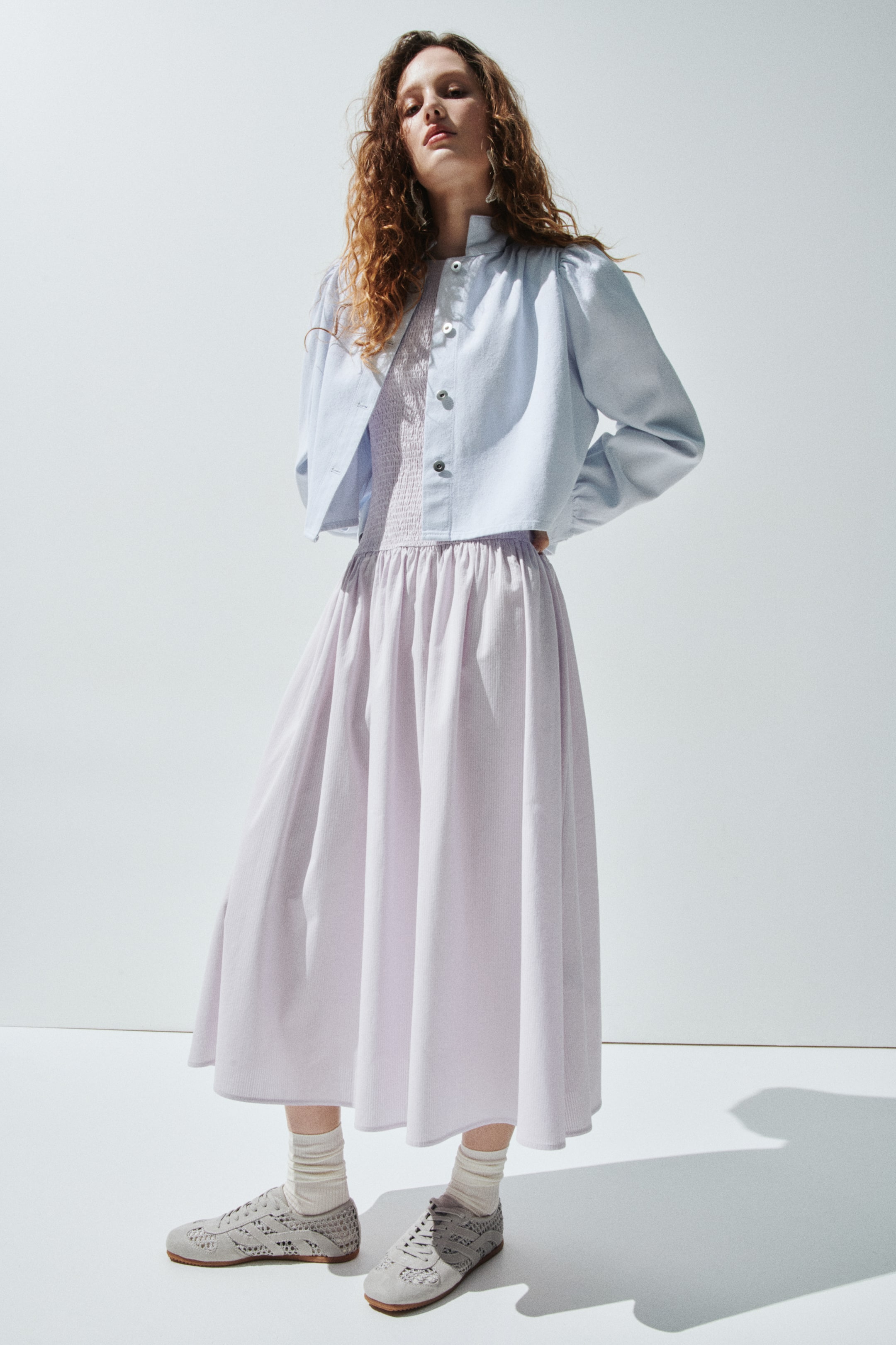 הצגת תמונה גדולה יותר: ז'קט קצר בצבע כחול בהיר, עם צווארון בנד, כתפיים תפוחות ושרוולים מקובצים, נלבש פתוח מעל שמלת מידי בצבע סגול בהיר. השמלה מציגה חזית מעוצבת בסגנון סמוק וחצאית זורמת ומקובצת. נעלי סניקרס אפורות עם חורים נלבשות עם גרביים בצבע קרם עם דוגמת צלעות.