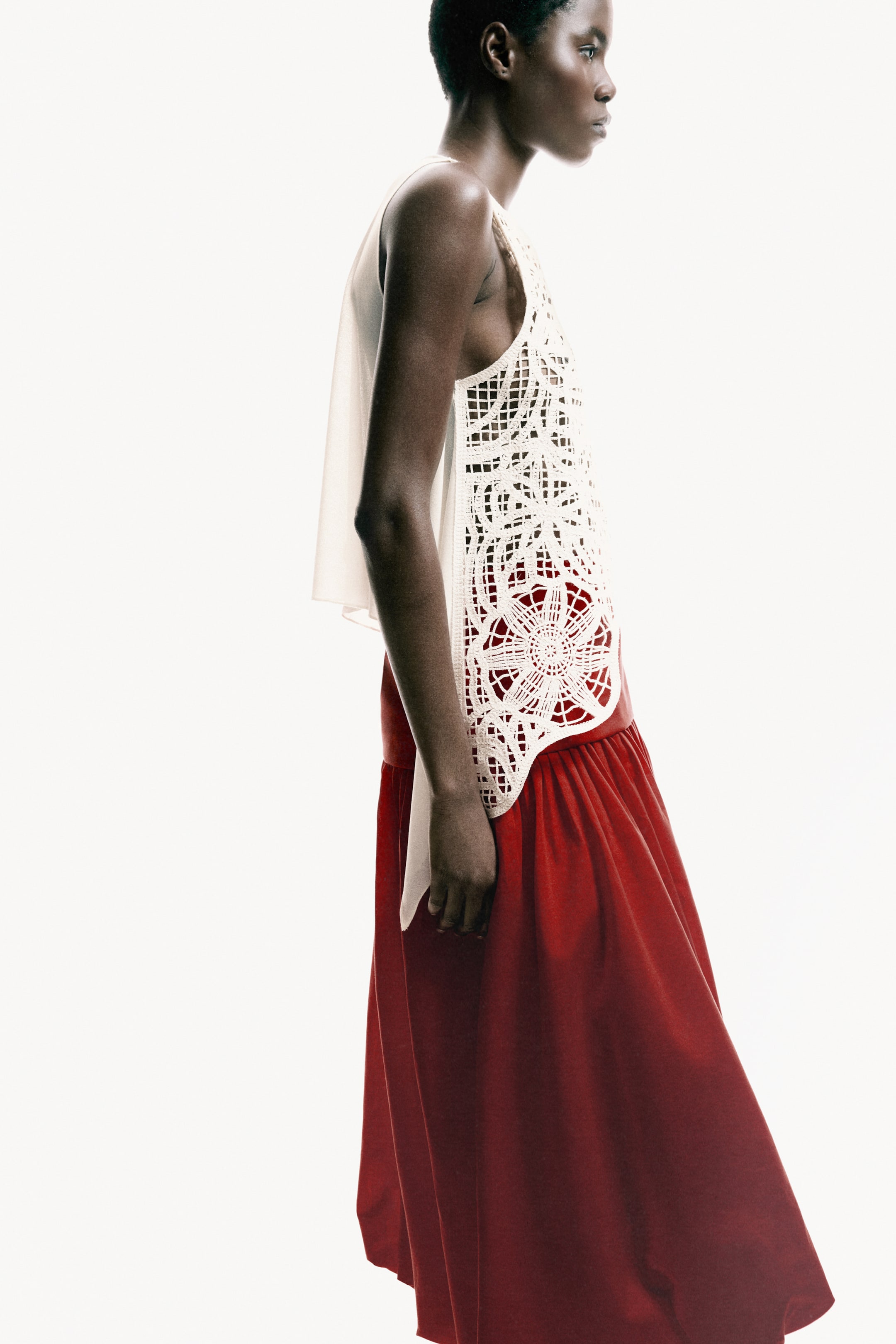 View larger image: A sleeveless, cream-colored top combines an open-knit, floral-patterned front panel with a sheer, flowing side overlay. This top is worn over a red, gathered maxi skirt, shown from a side profile, with the skirt's color visible through the top's open-knit design.