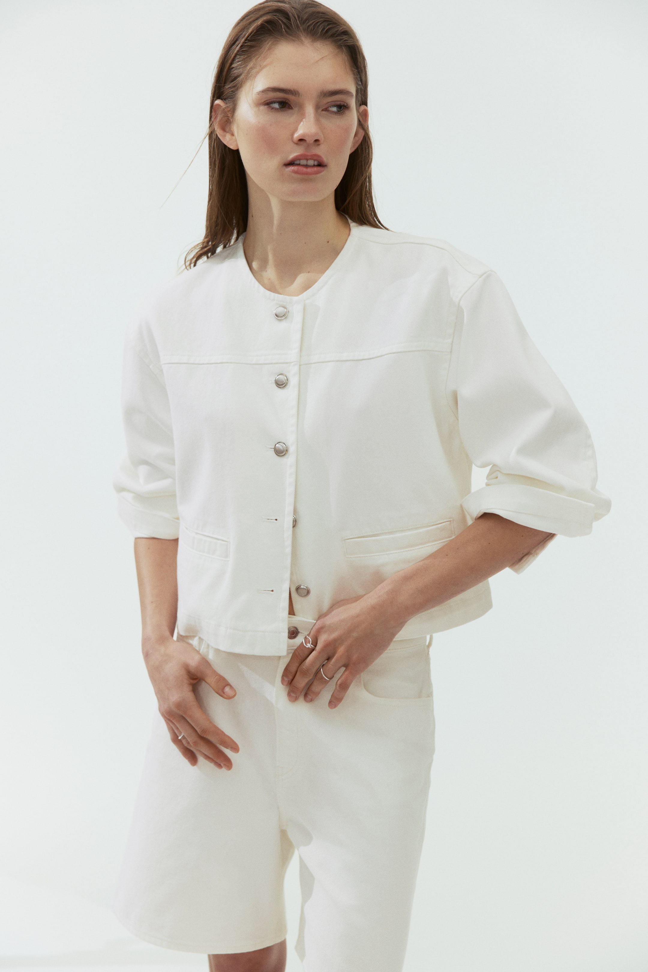 View larger image: A white, cropped, collarless jacket features silver-toned snap buttons down the front, horizontal chest seams and two patch pockets, with the long sleeves rolled up. Matching white shorts are also visible, sitting above the knee.