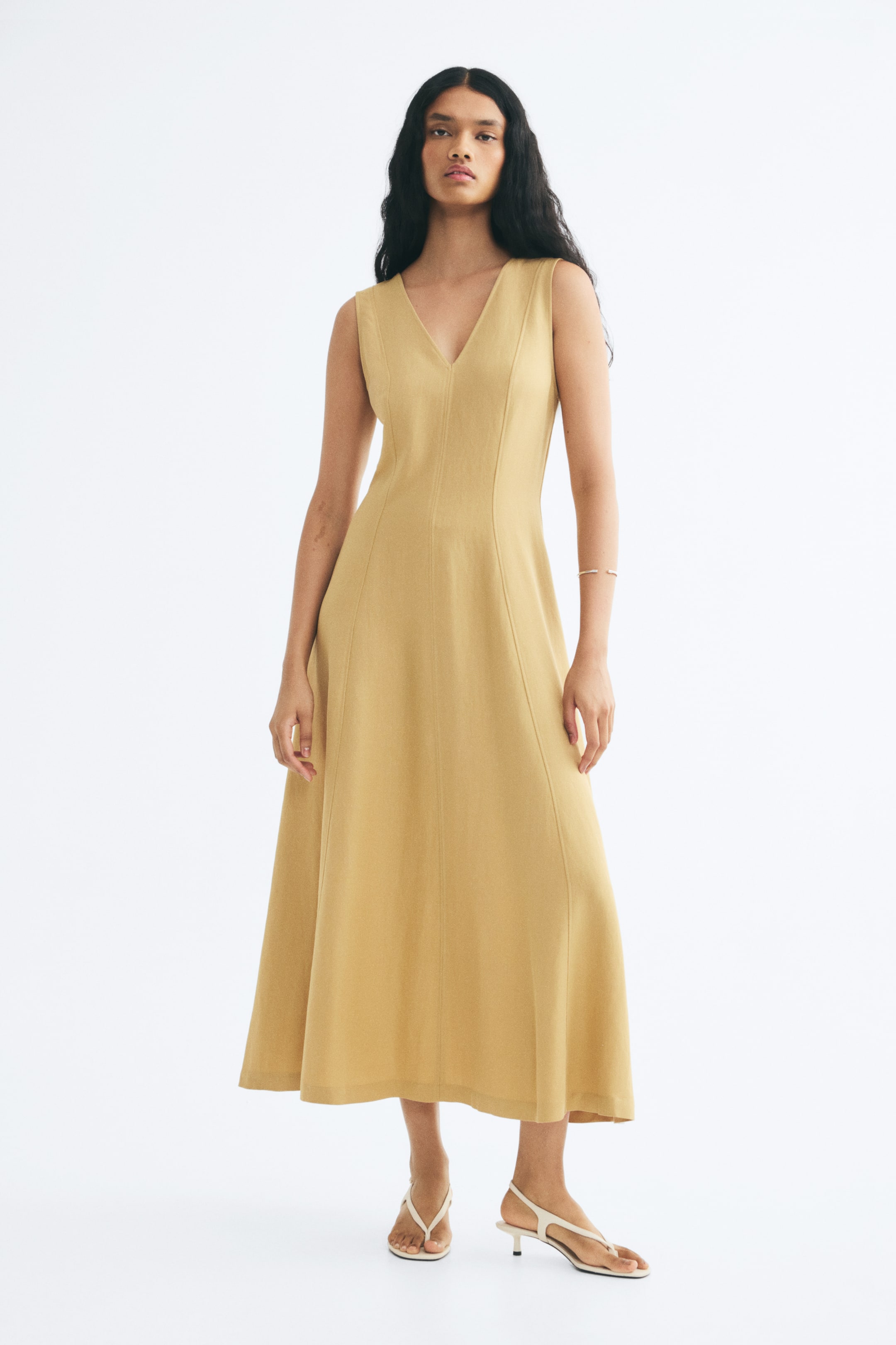 View larger image: A light yellow, sleeveless, calf-length dress is being worn by the model, who is standing facing forward. The dress features a V-neck, a fitted bodice, visible vertical seams running down the front, and a flared skirt.