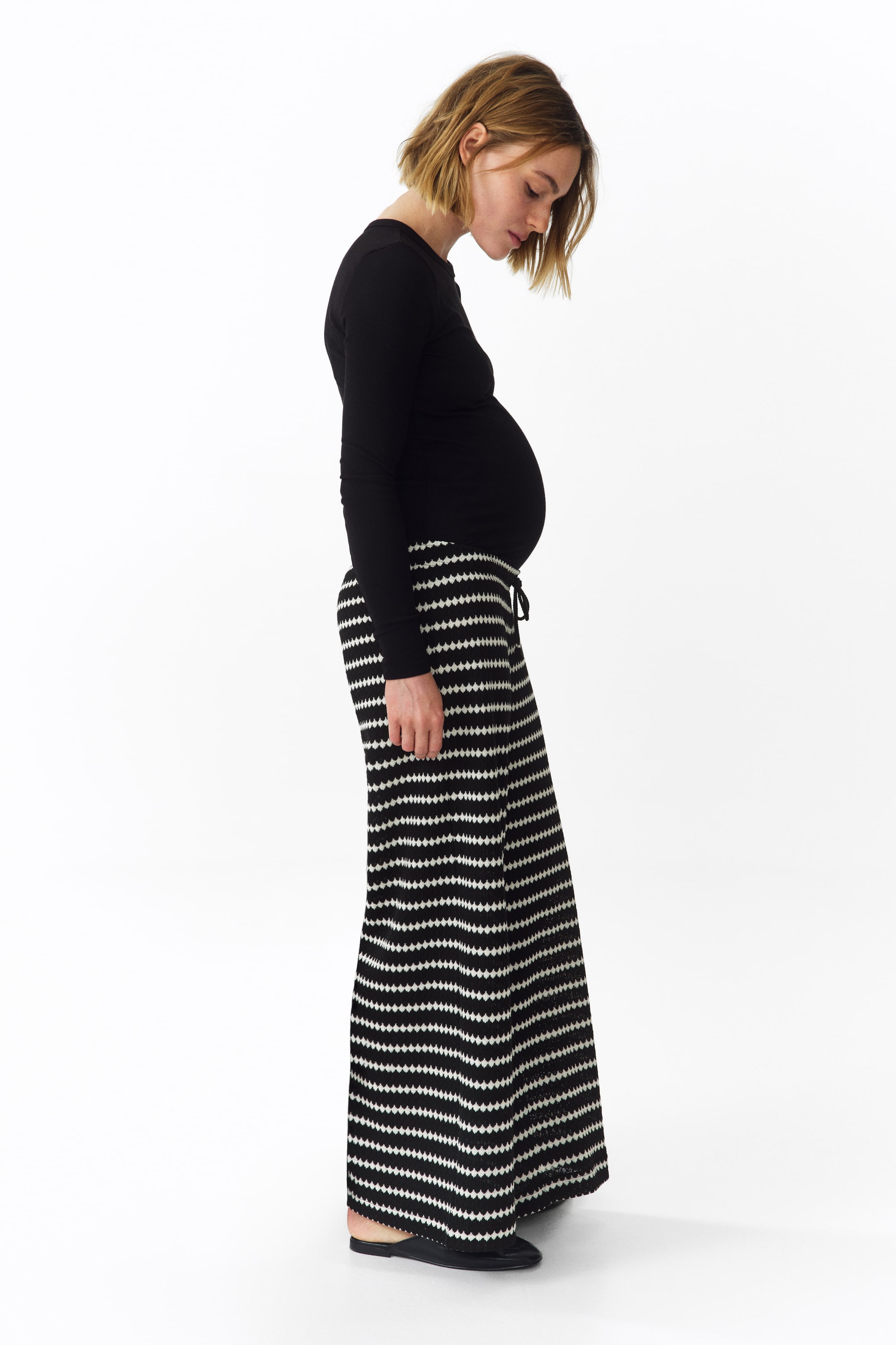 Grotere afbeelding bekijken: Een zwart top met lange mouwen en ronde hals rekt zich uit over de buik, gedragen met zwart-wit horizontaal gestreepte wijde broek met een trekkoord in de taille, gepresenteerd vanuit een zijaanzicht.