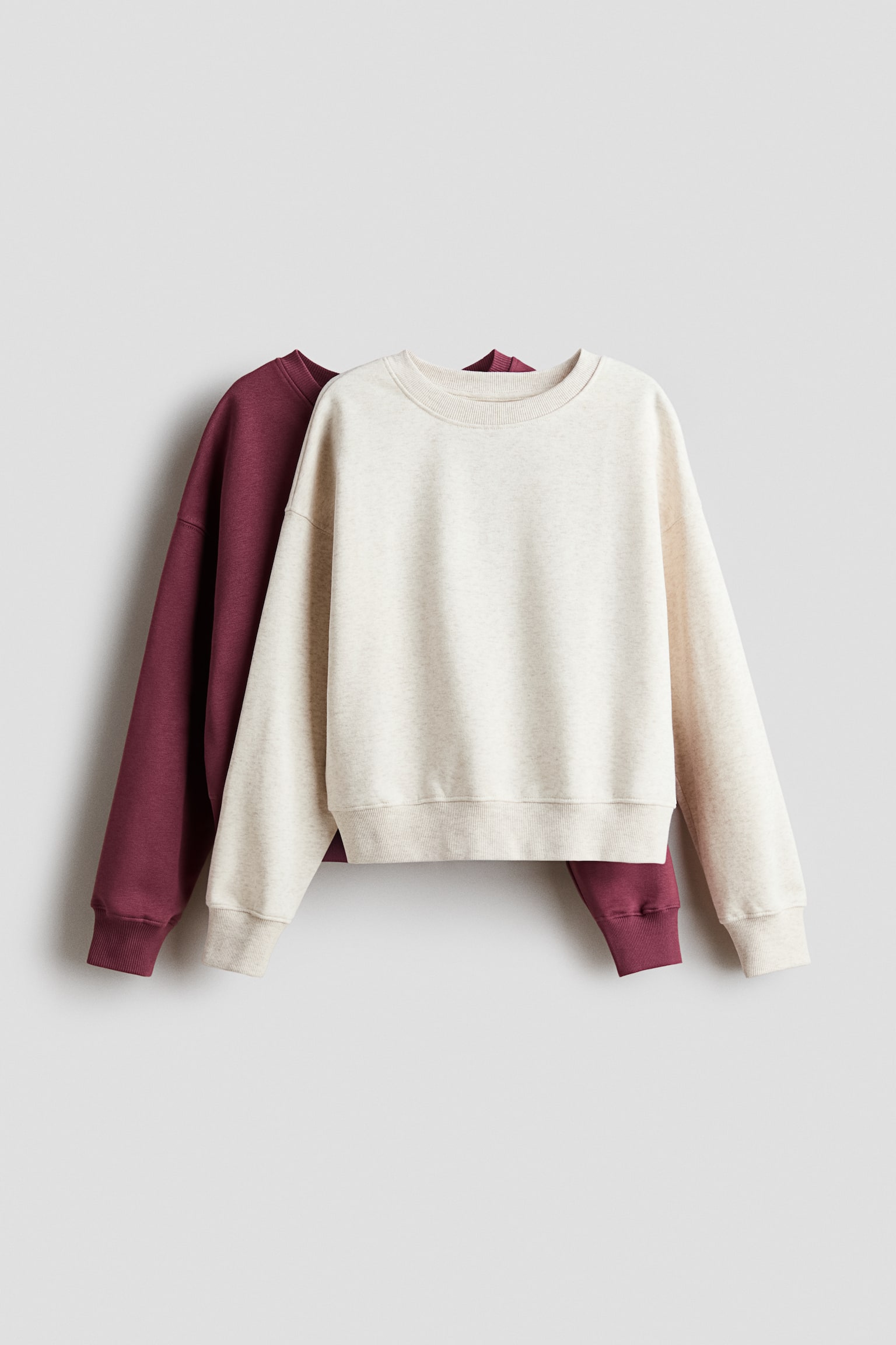 To løstsiddende sweatere i børstet fleece hænger frontvendt, med en lys havregrynsfarvet sweater lagt direkte foran en bordeauxfarvet. Begge sweatere har en rund hals med ribkant, sænkede skuldre samt ribkanter ved manchetter og søm.