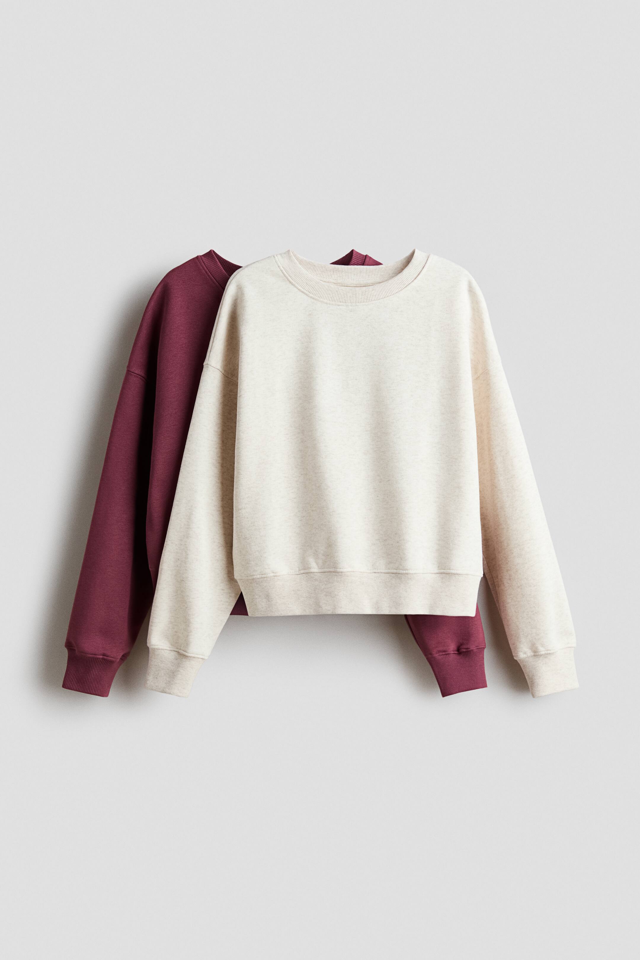 View larger image: Two loose-fit sweaters in brushed fleece fabric are hanging front-facing, with a light oatmeal-coloured sweater layered directly in front of a burgundy one. Both sweaters feature a round, rib-trimmed neckline, dropped shoulders, and ribbing at the cuffs and hem.