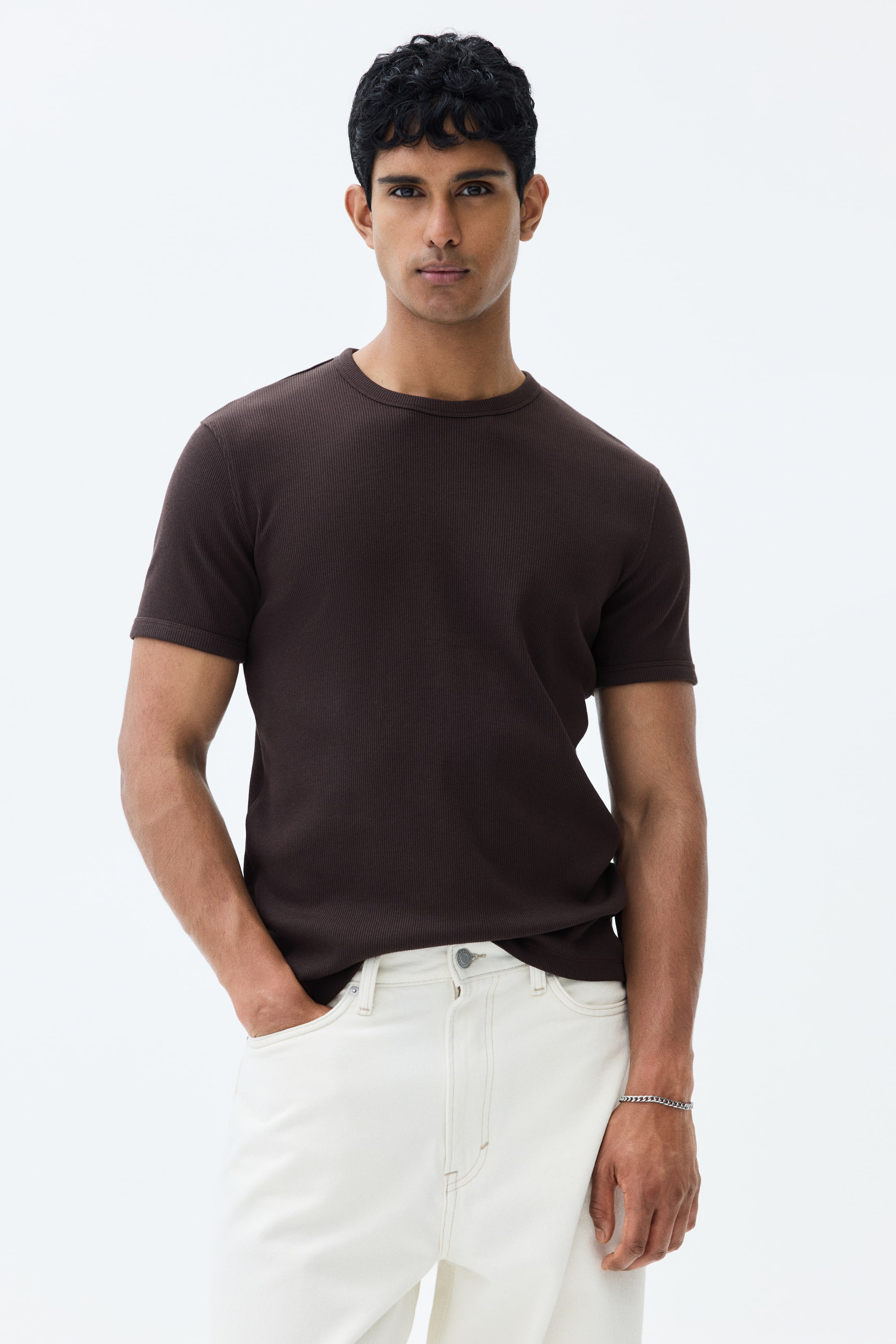 View larger image: The model is wearing a dark brown ribbed cotton jersey T-shirt. It comes with a round neckline, short sleeves, and a straight-cut hem. The slim fit accentuates the model's body contours.